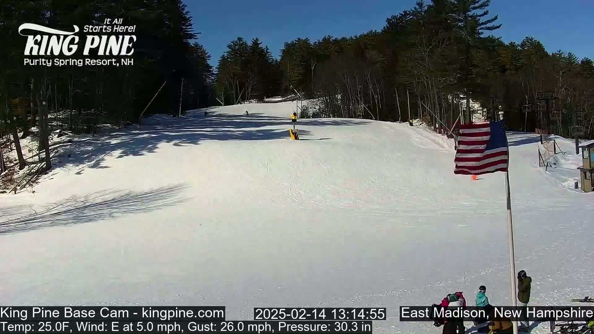 A snow-covered ski slope on a sunny day with several skiers visible, surrounded by a treeline.