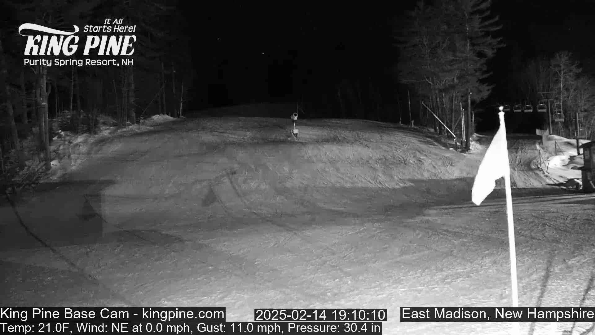 A nighttime view of a snow-covered ski slope with a chairlift and flag in the background.