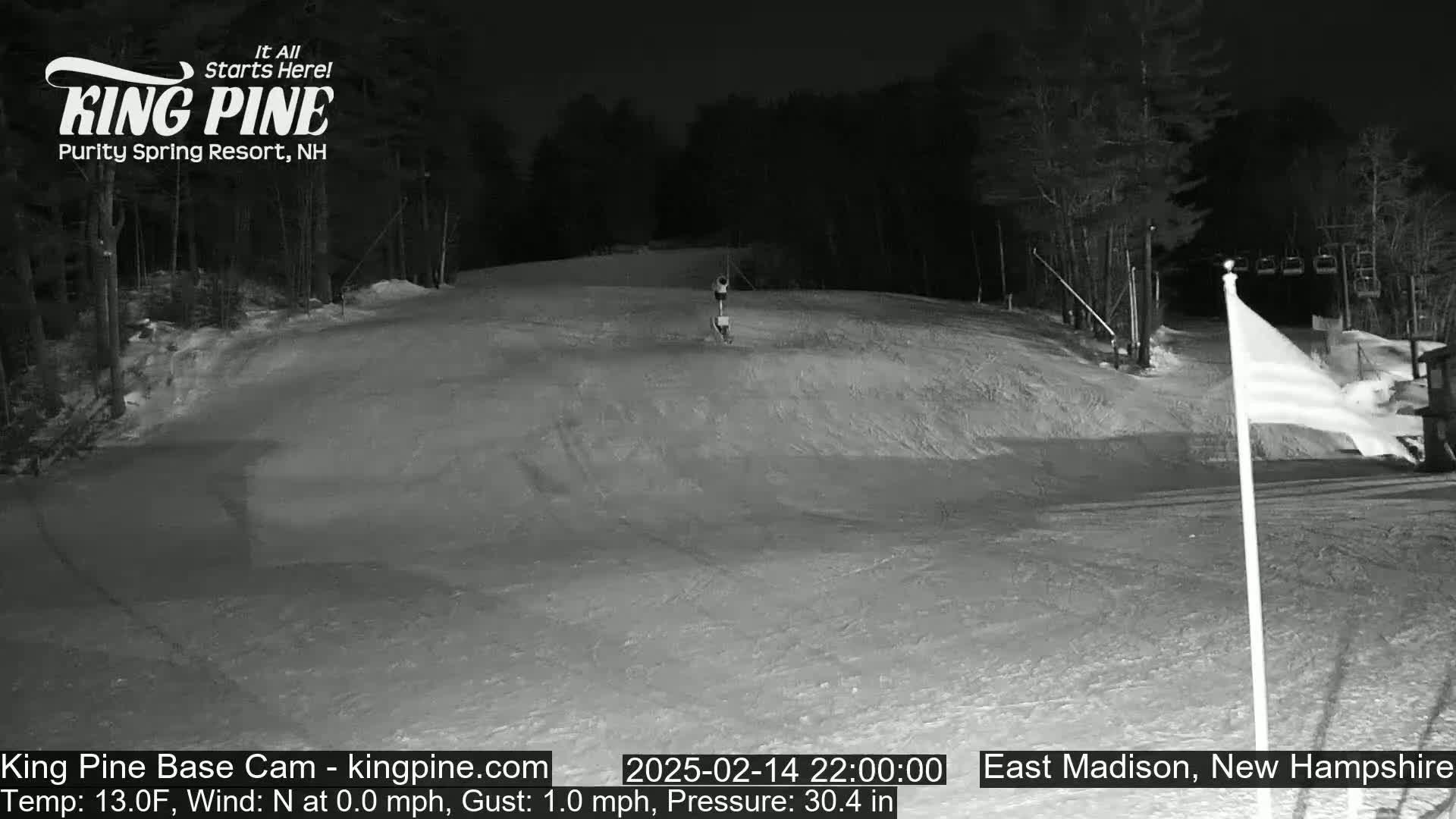 A snow-covered ski slope at night, illuminated by artificial light, with a lone figure visible on the slope and a chairlift in the background.