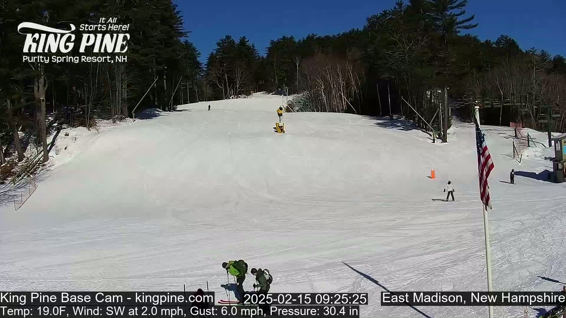 A snow-covered ski slope with several skiers and snowboarders on it, under a clear blue sky.