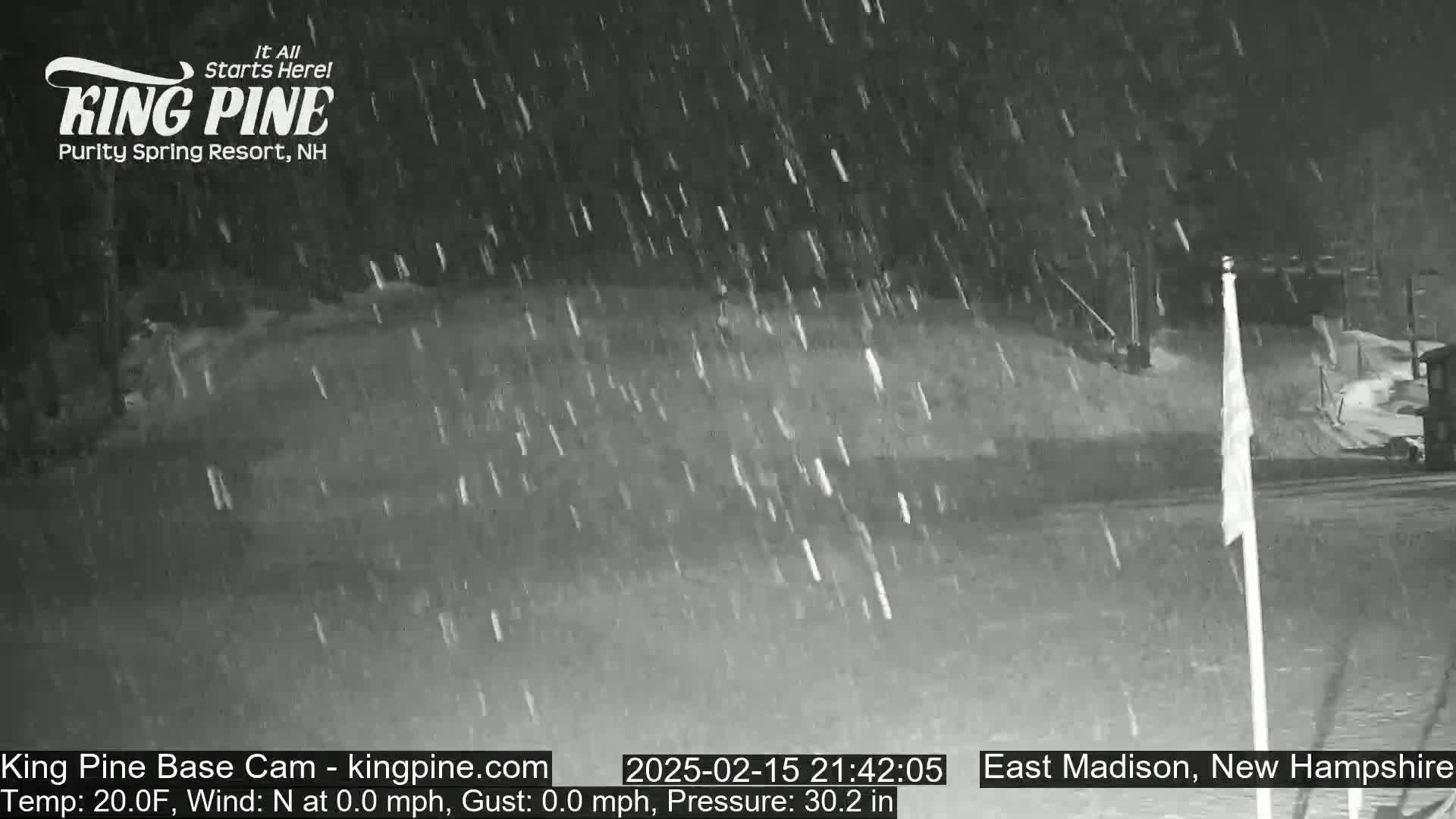 A nighttime snowstorm blankets a snowy landscape with a flagpole visible in the foreground.