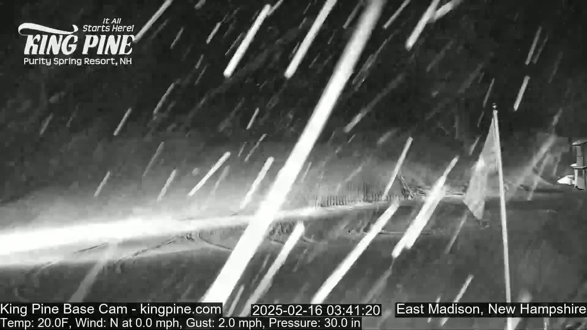 Heavy snowfall at night obscures a mostly-flat, snow-covered landscape with a tall flagpole and a building partially visible in the distance.