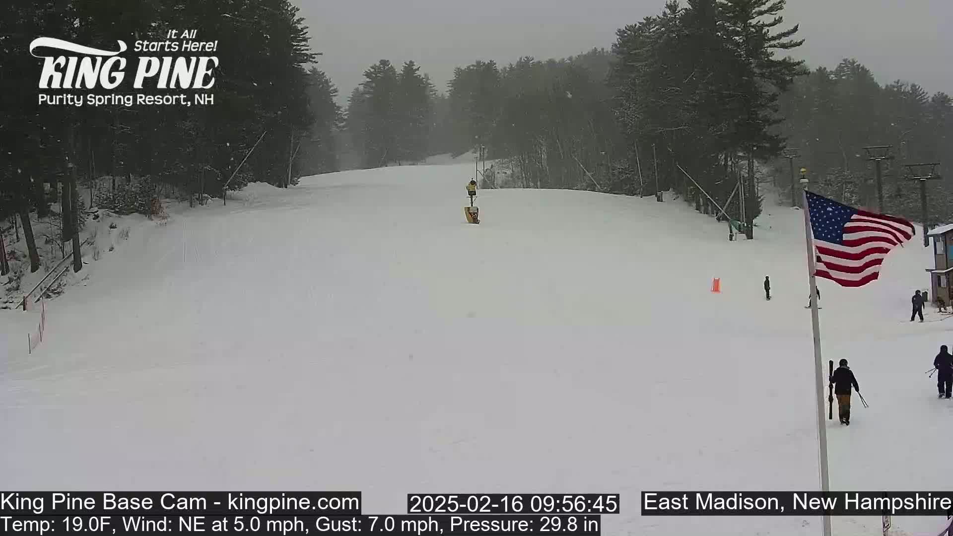A snow-covered ski slope with a few skiers and a US flag is visible on a cloudy day with a northeast wind.