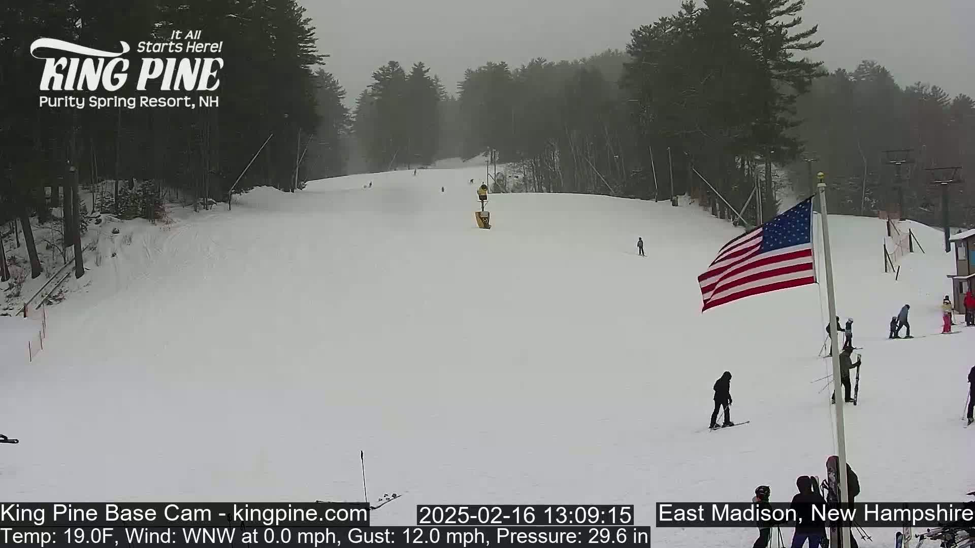 A snowy ski slope with several skiers and snowboarders visible, under an overcast sky.