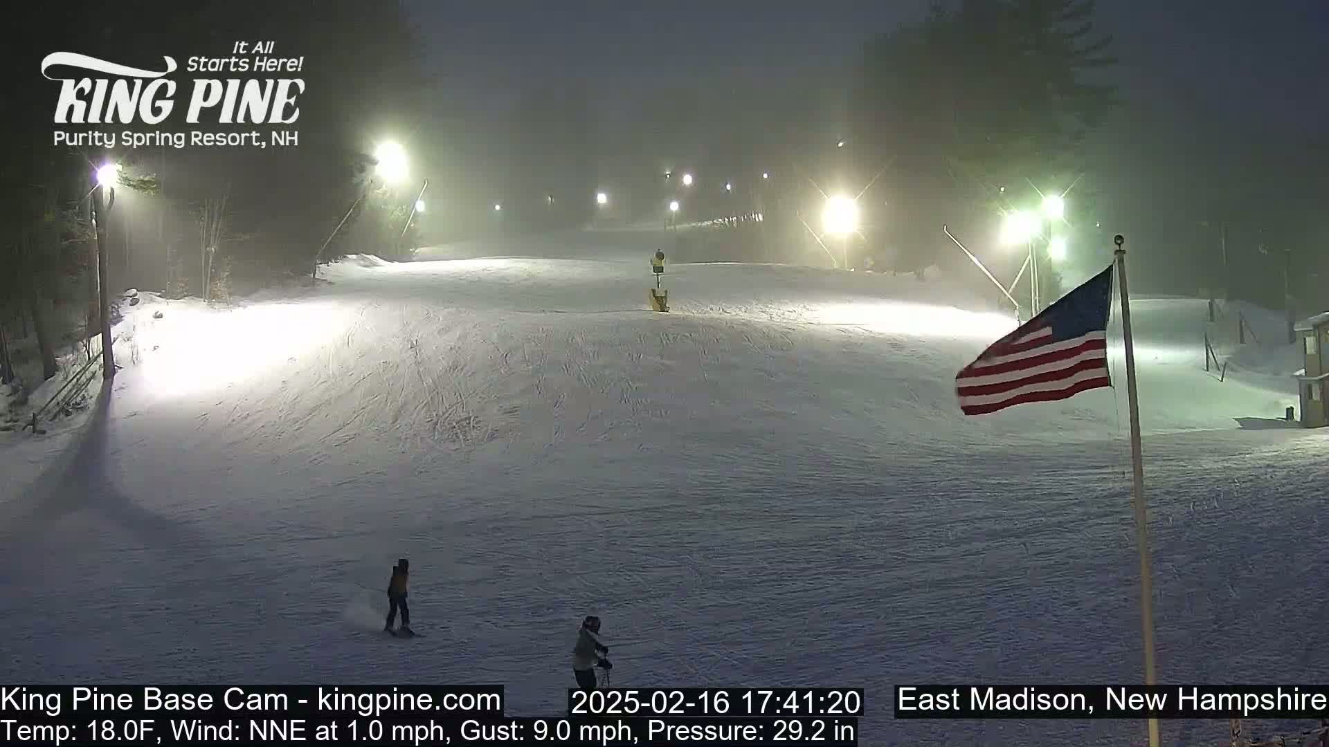 A nighttime view of a snow-covered ski slope with a person snowboarding, illuminated by lights, under hazy conditions.