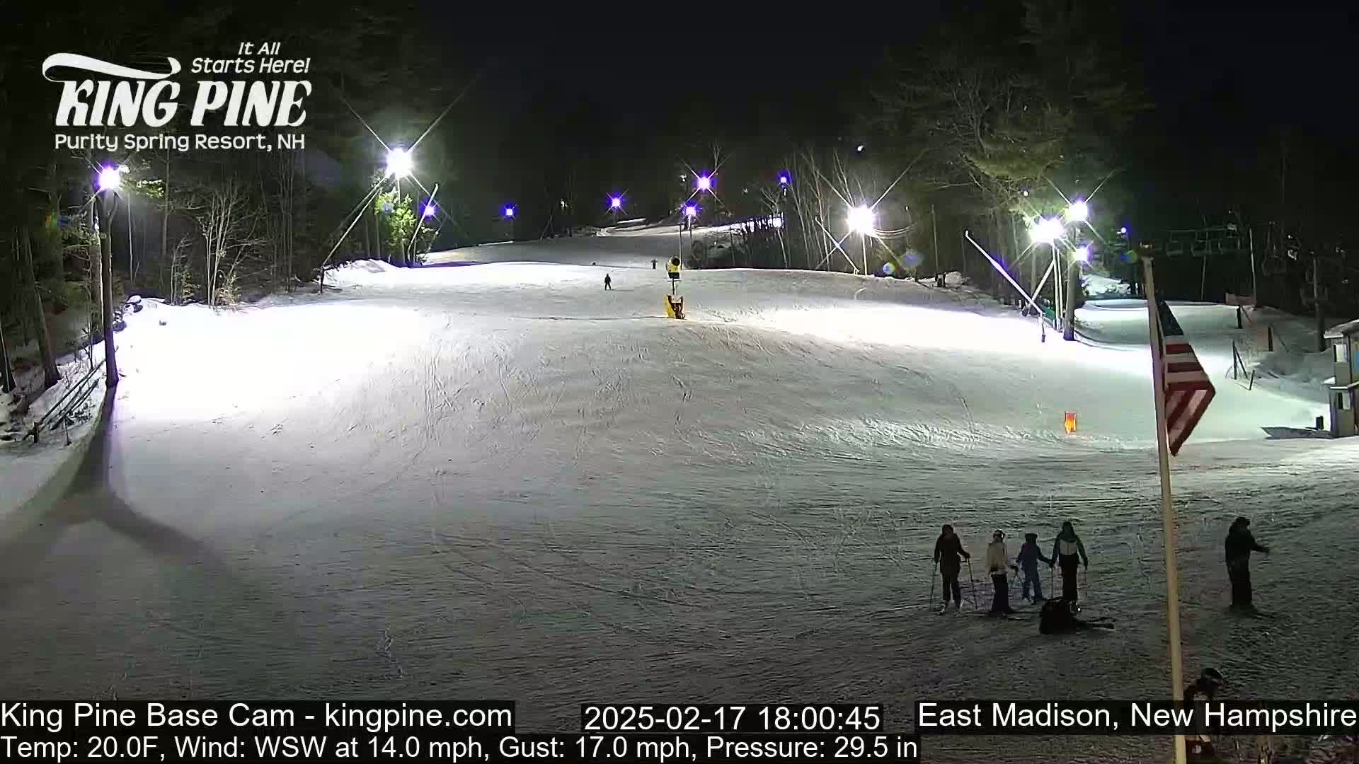 A nighttime view of a snow-covered ski slope illuminated by artificial lights, with several skiers visible at the bottom of the hill and a flagpole with an American flag in the foreground.