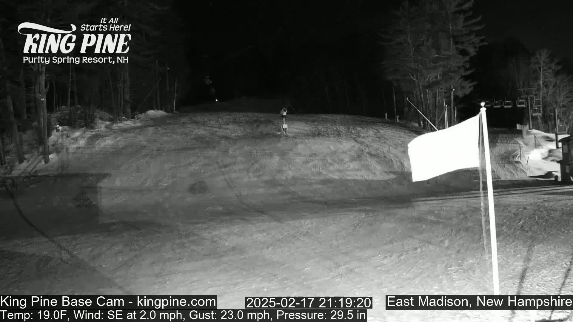 A snow-covered ski slope at night is illuminated, with a white flag waving in a southeast wind under a dark sky.