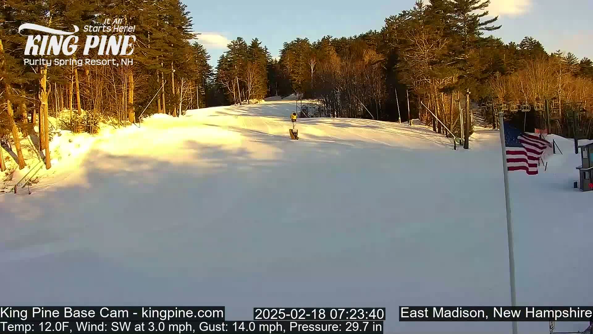 A snow-covered ski slope is visible under a clear, bright sky, with trees lining the background and a small American flag in the lower right.