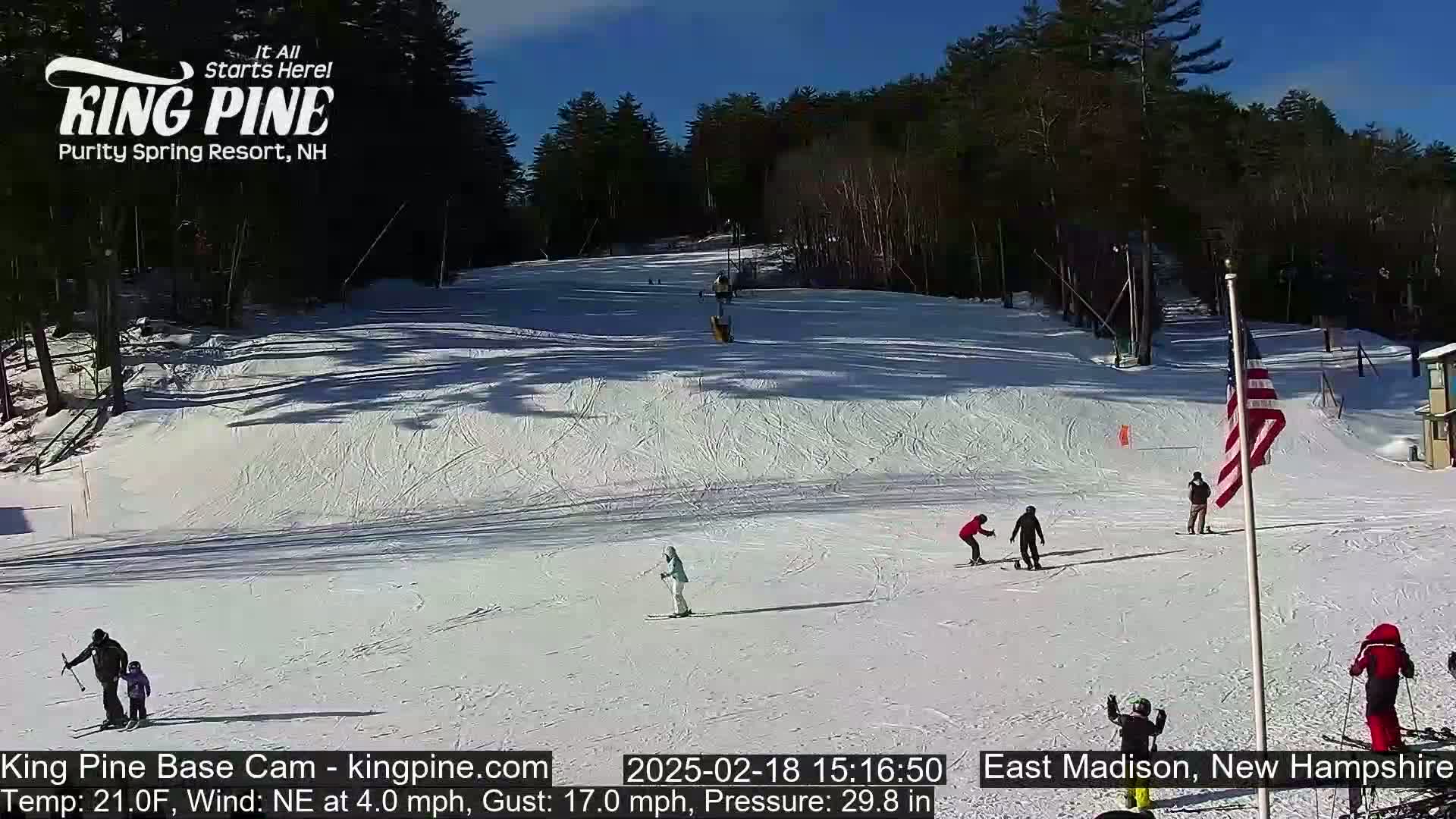 Several skiers and snowboarders enjoy a sunny, snow-covered ski slope on a clear day with a northeast wind.