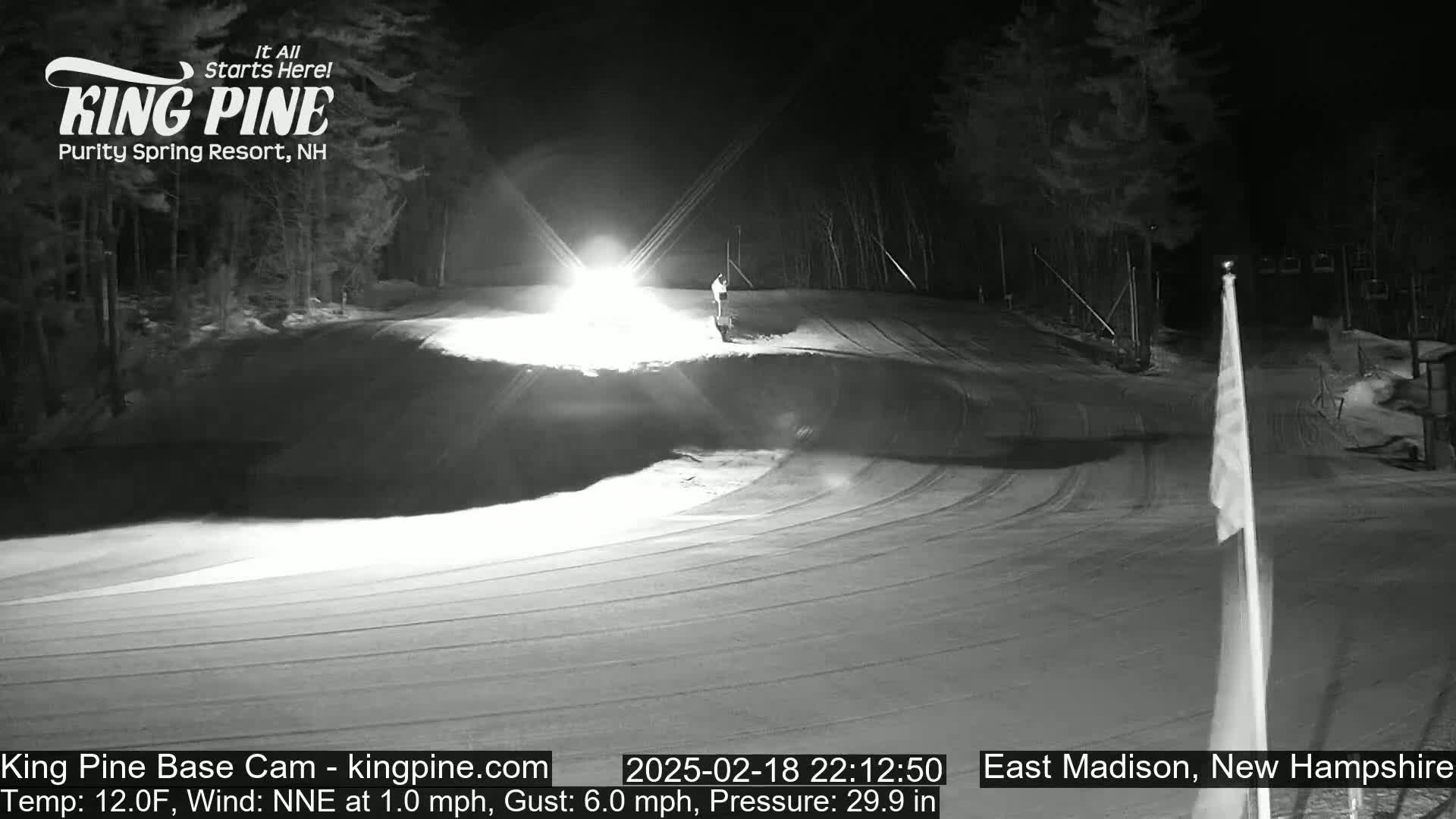 A nighttime, snow-covered ski slope is illuminated by a bright light, with a lone figure visible near the top and a flagpole standing to the right.