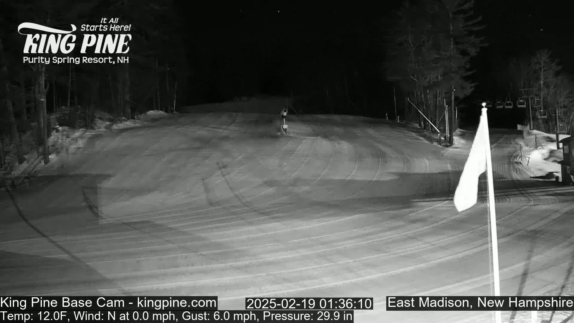 A nighttime, snow-covered ski slope with a person and a chairlift visible in the distance under a clear sky.