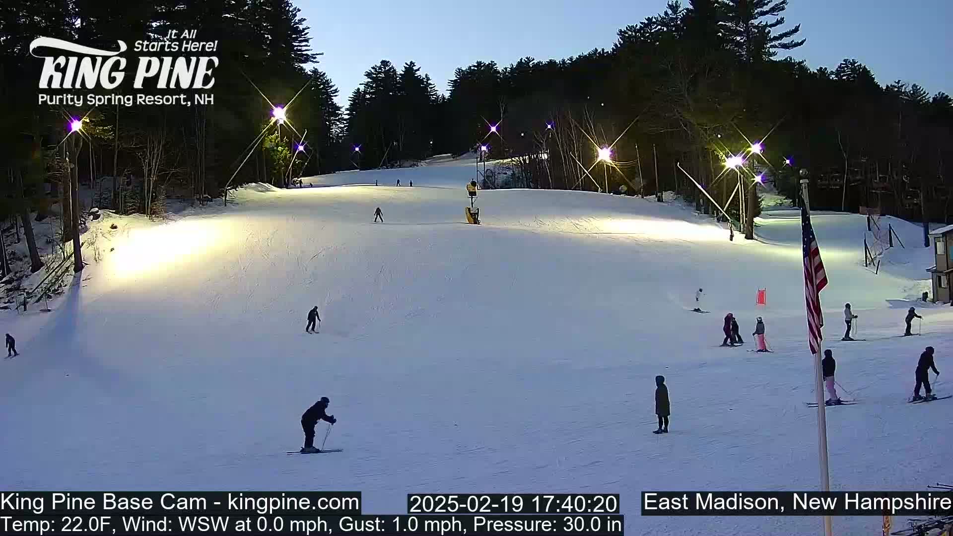 A snow-covered ski slope at night is illuminated by several lights, with several skiers and snowboarders visible on the slope under a twilight sky.