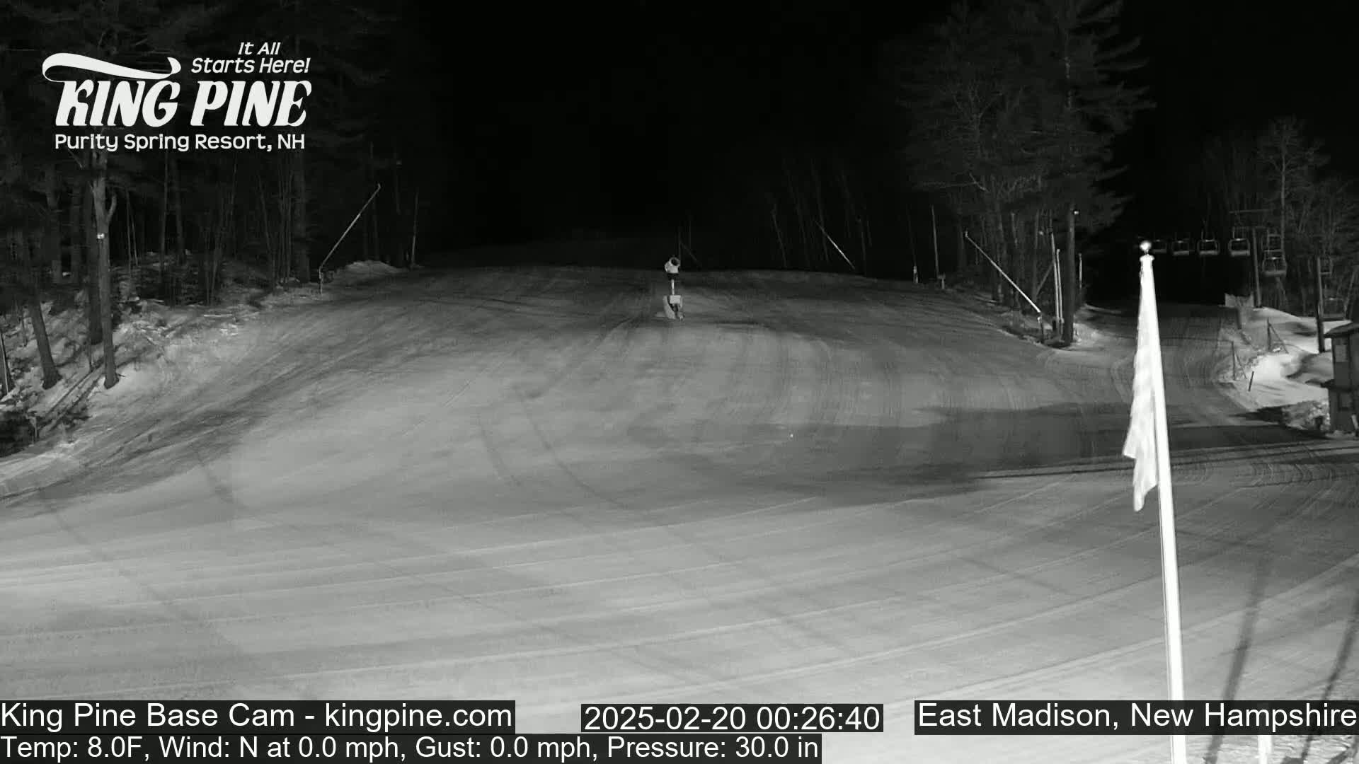 A snow-covered ski slope at night, with a chairlift visible in the background under a clear, dark sky.