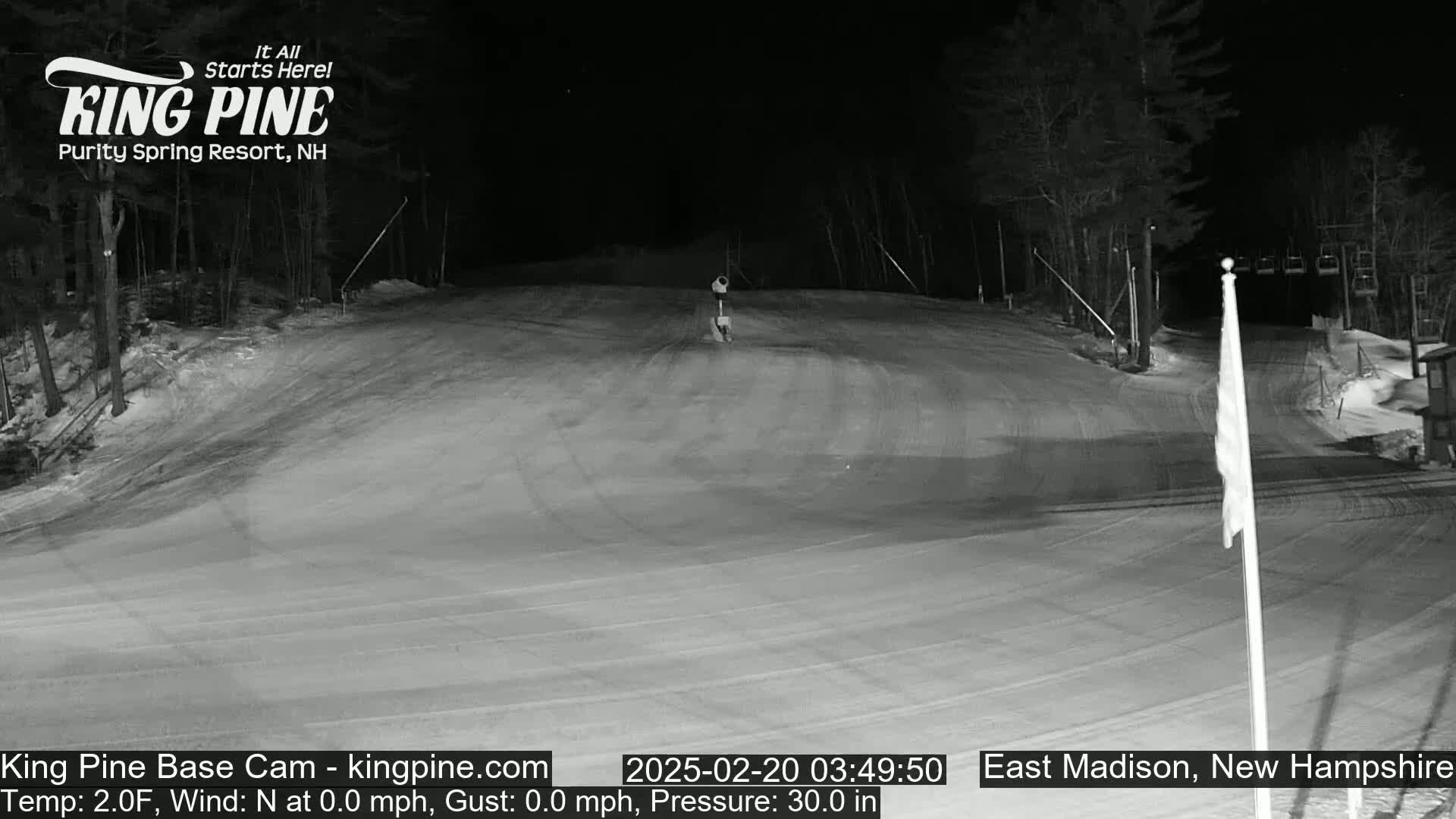 A snow-covered ski slope at night, illuminated by artificial light, with a ski lift visible in the background.