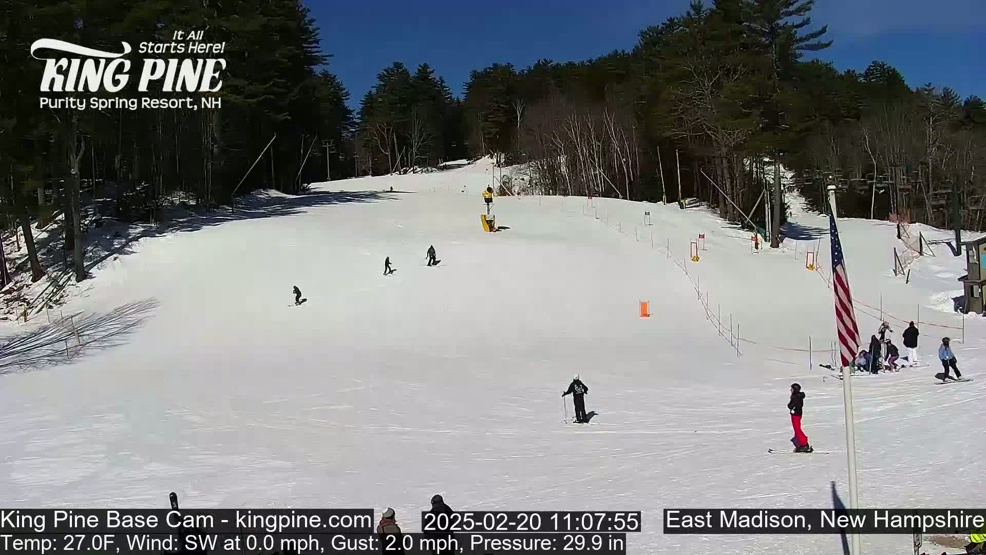 A sunny snow-covered ski slope with several skiers and snowboarders, surrounded by a wooded area.