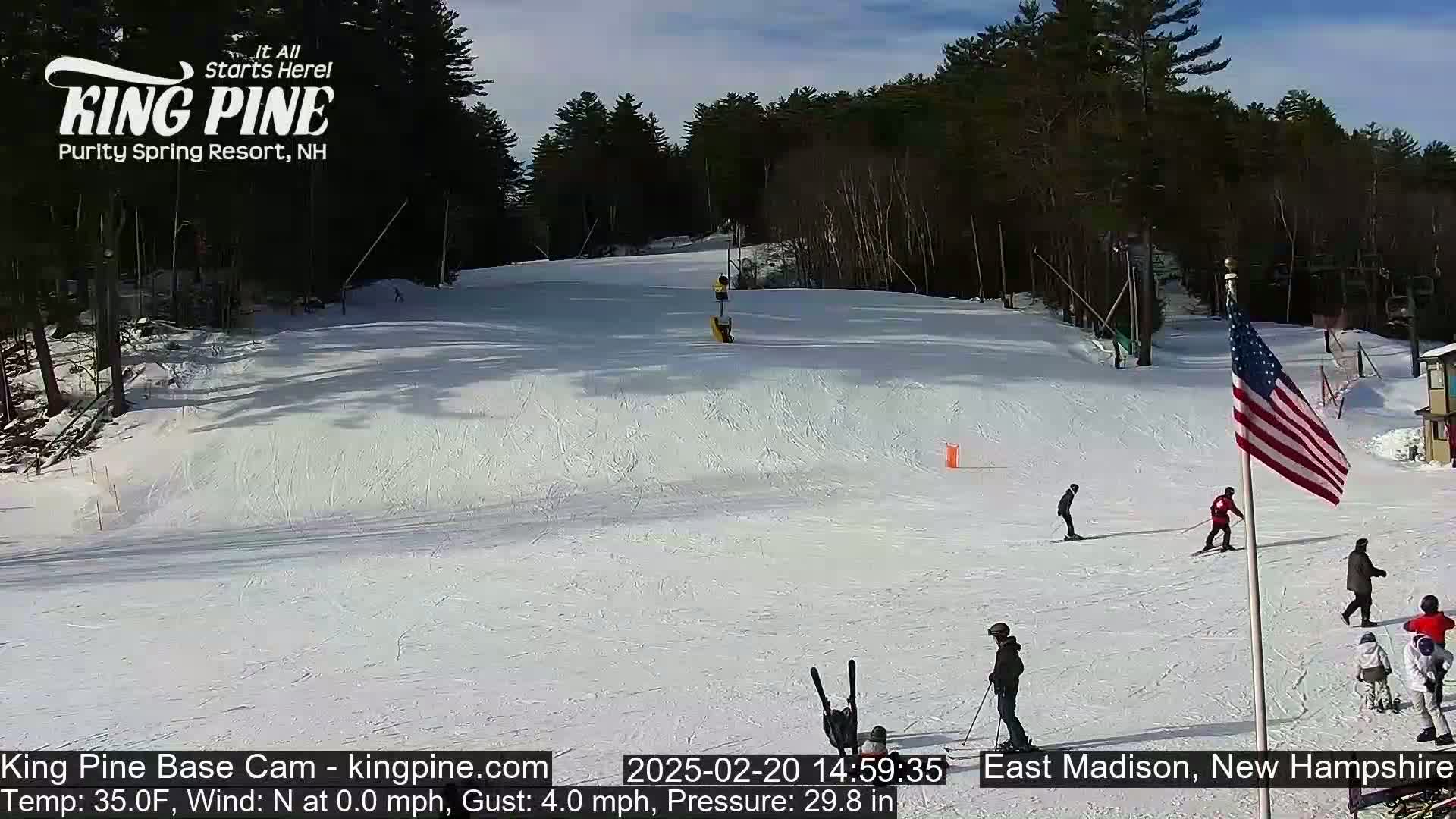 A sunny day at a snow-covered ski slope shows several skiers and snowboarders on a gently sloping hill surrounded by trees.