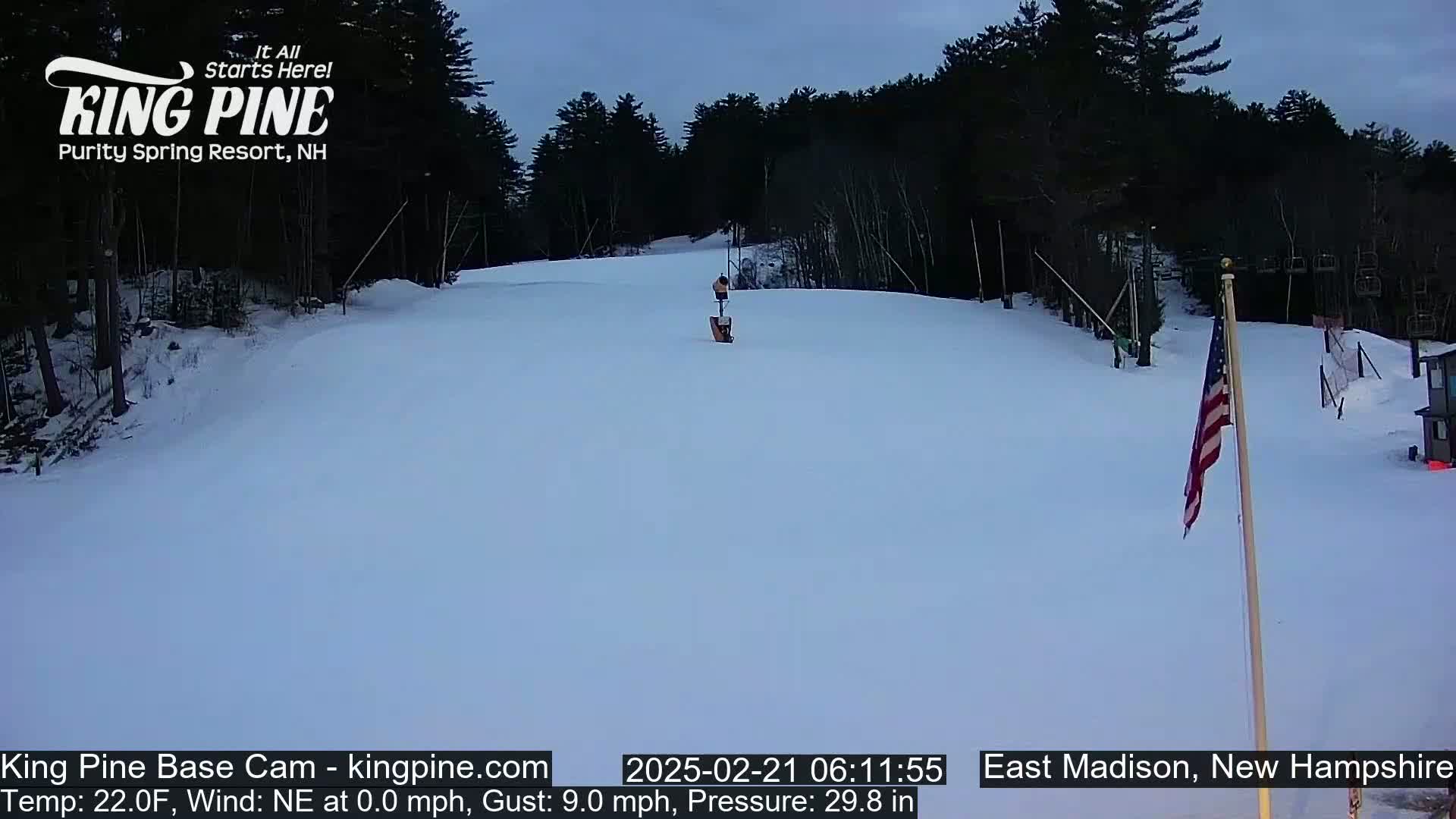 A snow-covered ski slope is seen under a twilight sky, with a lone figure and American flag visible in the distance.