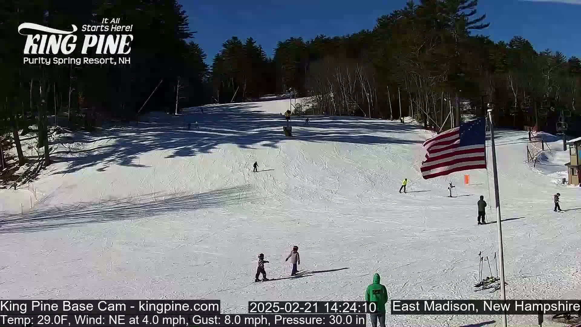 A sunny snow-covered ski slope with several skiers and snowboarders, a waving American flag, and a wooded backdrop.
