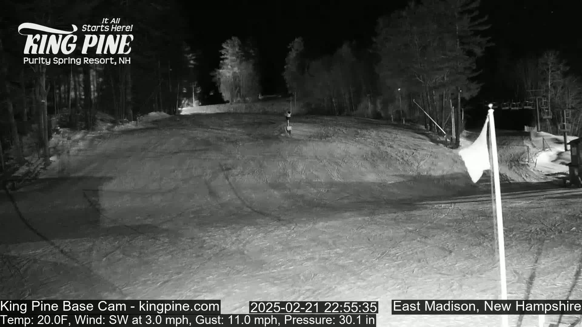 A nighttime, snow-covered ski slope is illuminated, with a lone figure visible near the center, under a clear sky and a southwest wind.