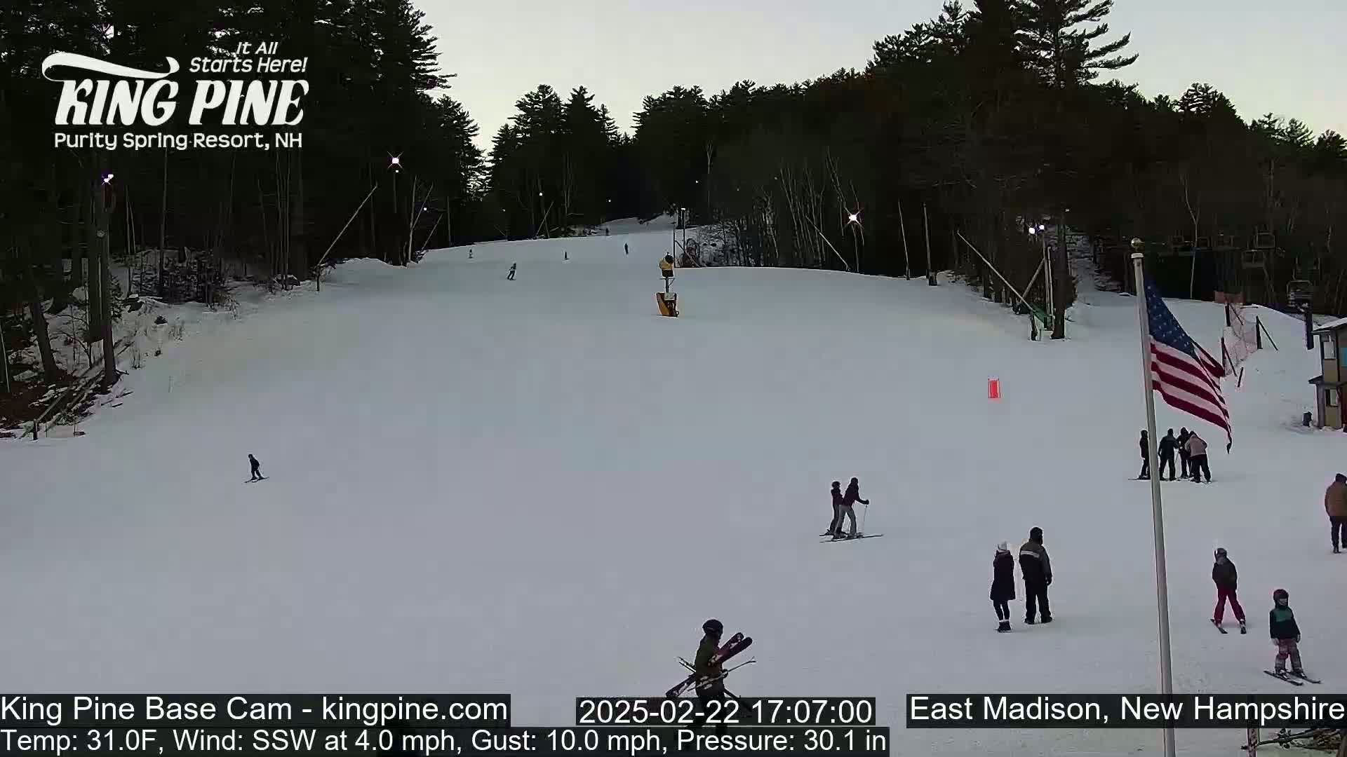 A snow-covered ski slope at dusk with several skiers and snowboarders, under a clear sky and a light south-southwesterly wind.