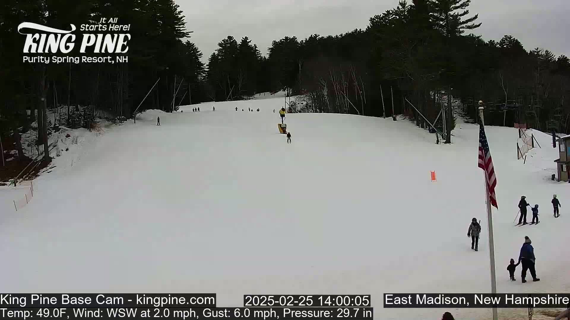 A snow-covered ski slope with several skiers and snowboarders, a chairlift, and trees in the background under an overcast sky.
