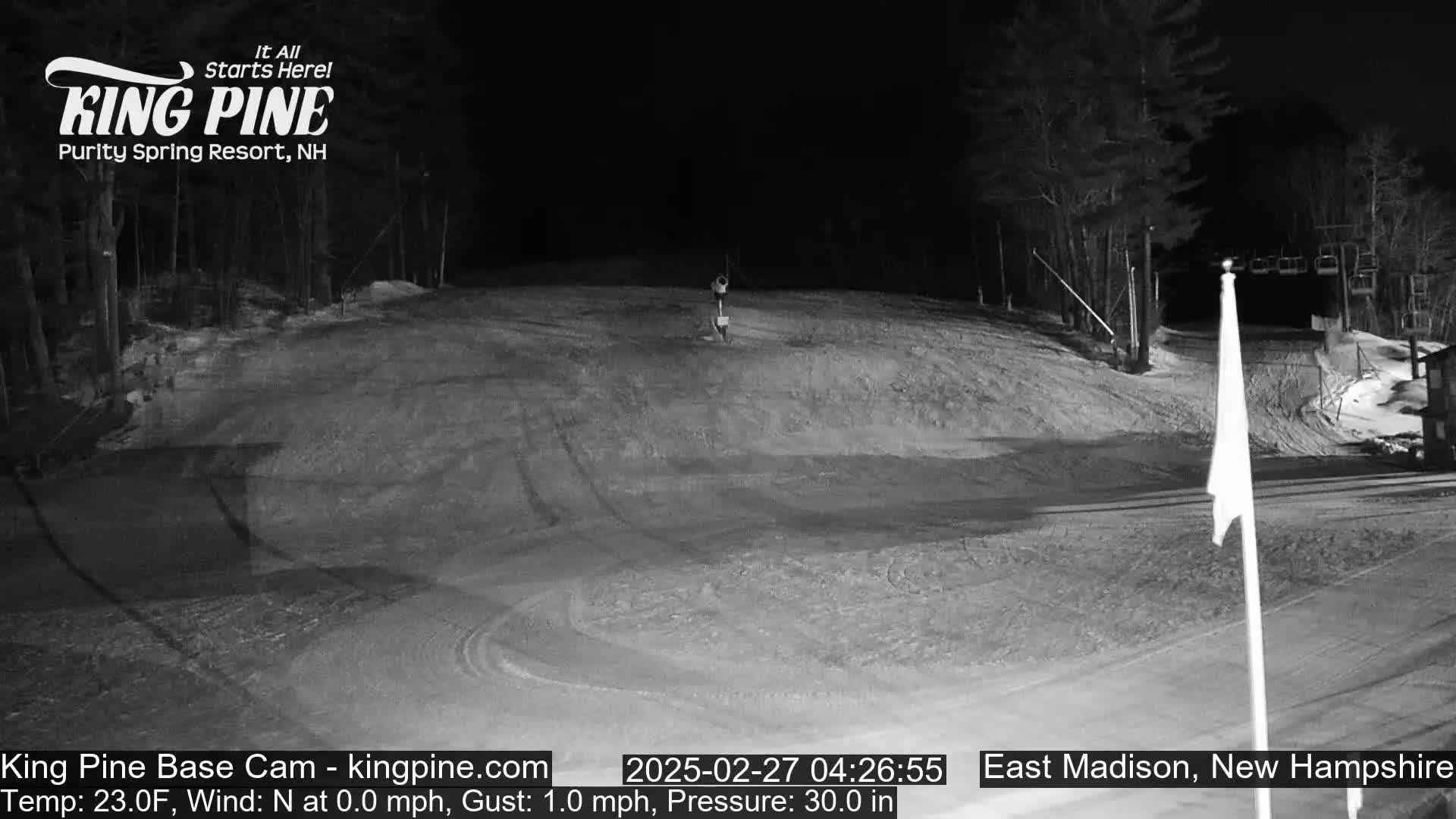 A snow-covered ski slope at night, illuminated by artificial light, with a ski lift visible in the background and a flagpole in the foreground.