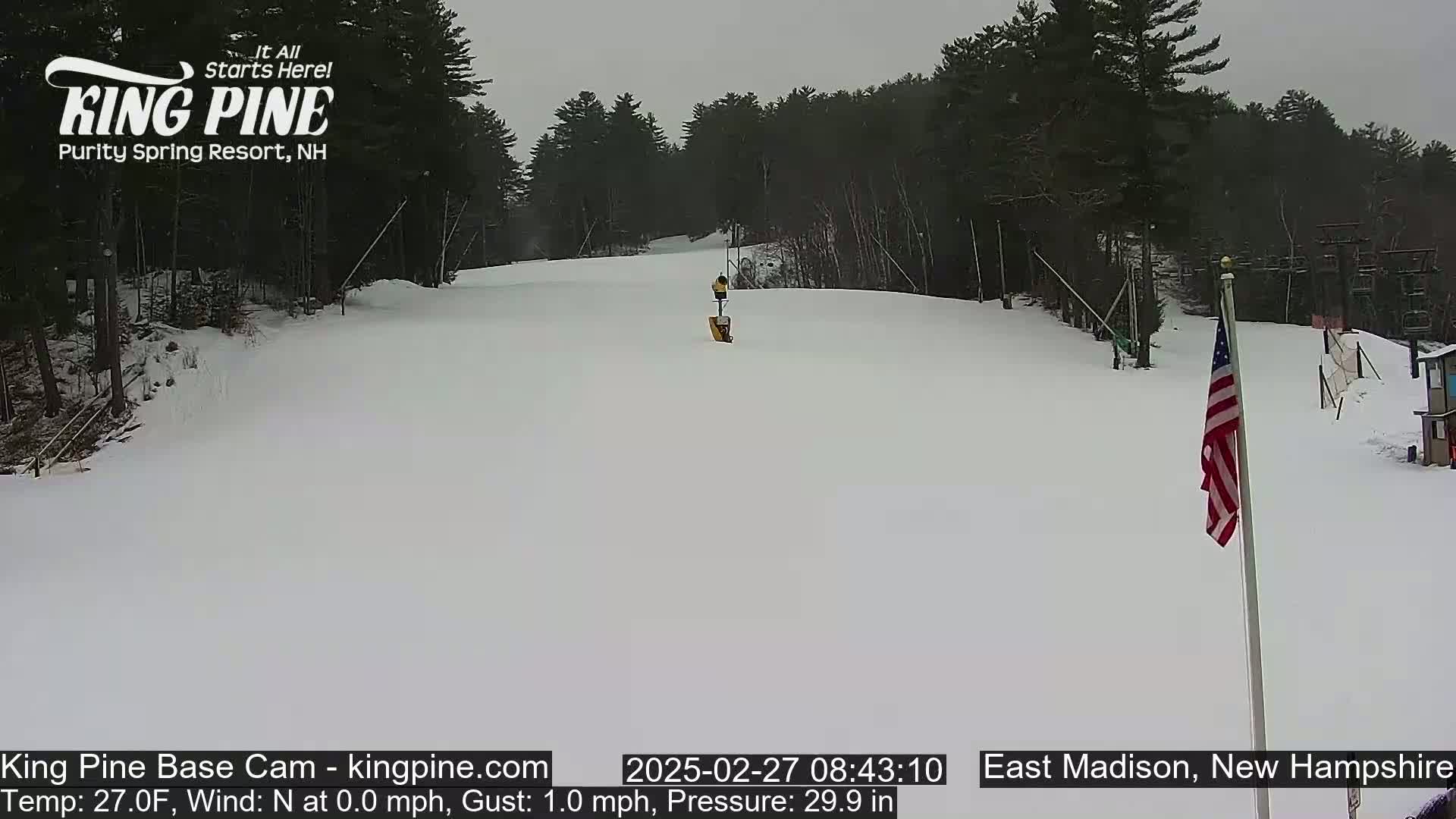 A snow-covered ski slope is visible, with a mostly cloudy sky and a lone figure in the distance near a snowmaking machine; an American flag stands to the right.