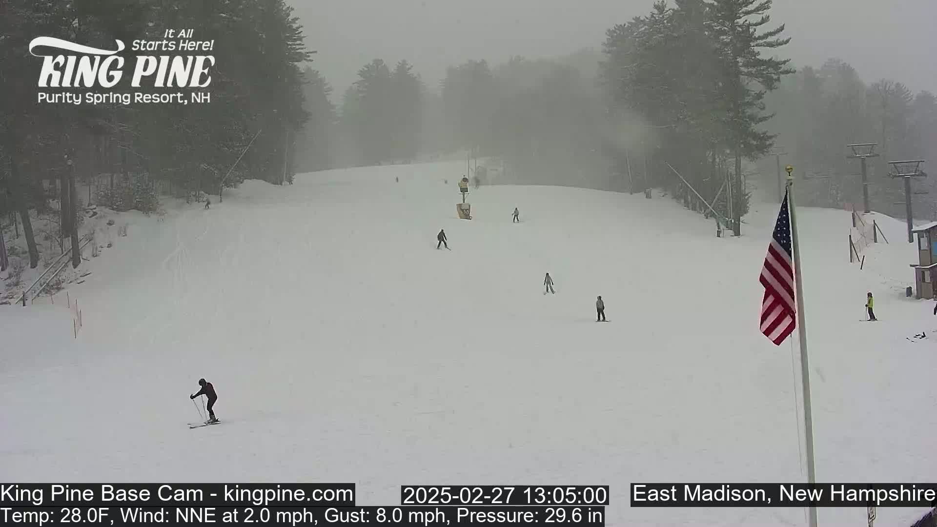 A snowy ski slope with several skiers and a US flag visible in the hazy, snowy weather.