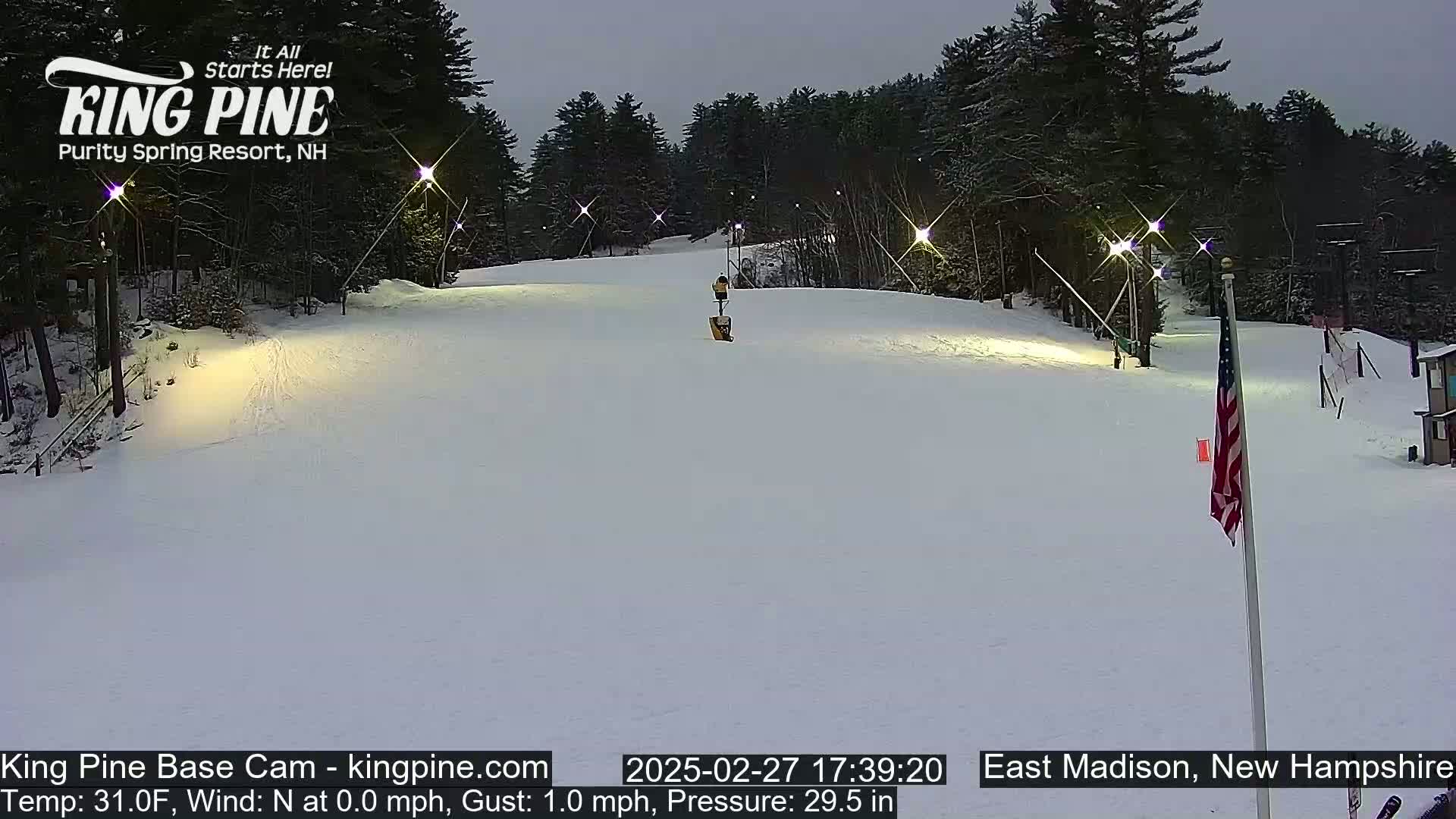 A snow-covered ski slope is illuminated at night by several lights, with a mostly clear, dark sky above and trees lining the background.