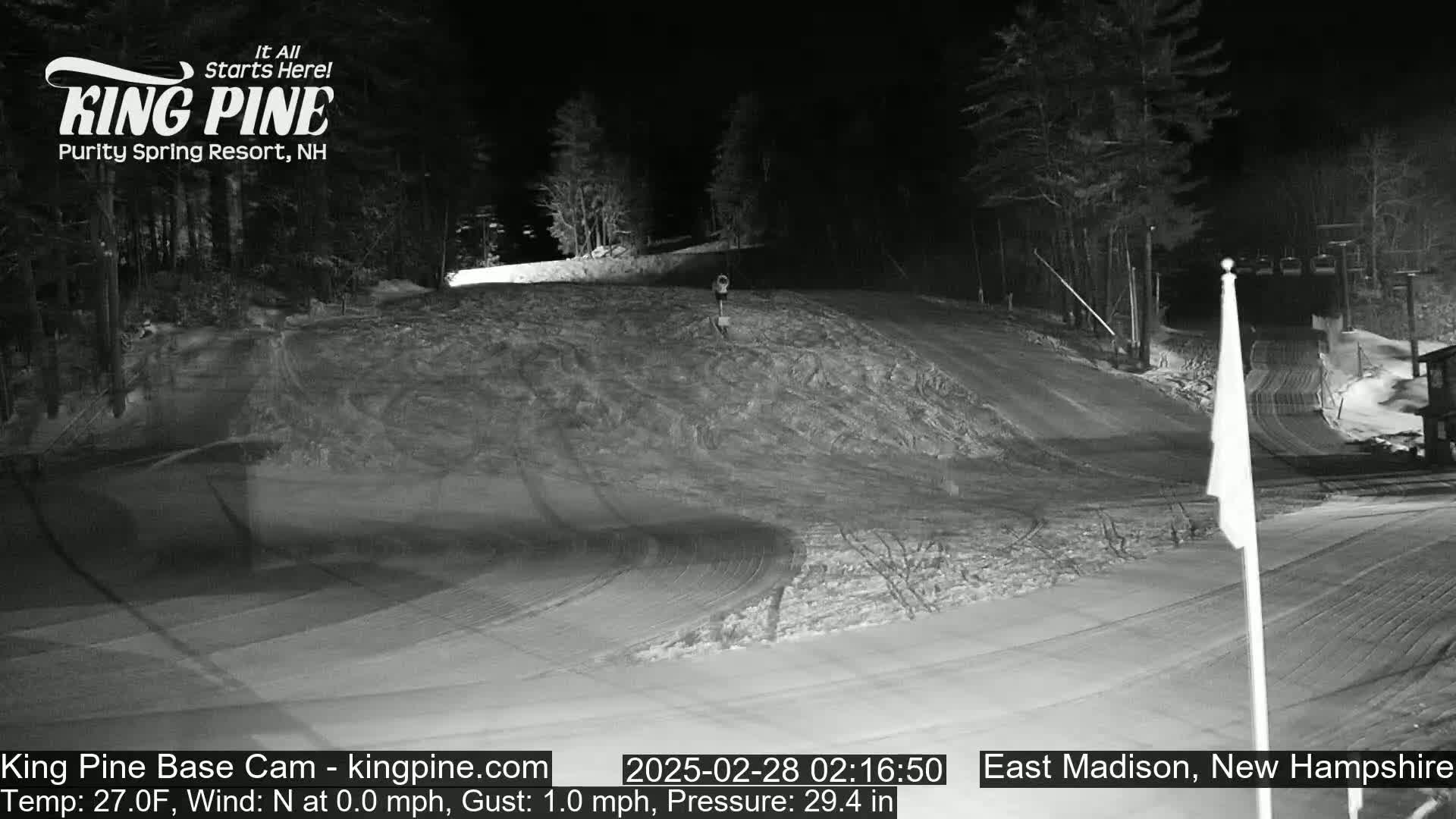 A snow-covered ski slope at night, illuminated by artificial light, with trees in the background and a calm, cold wind.