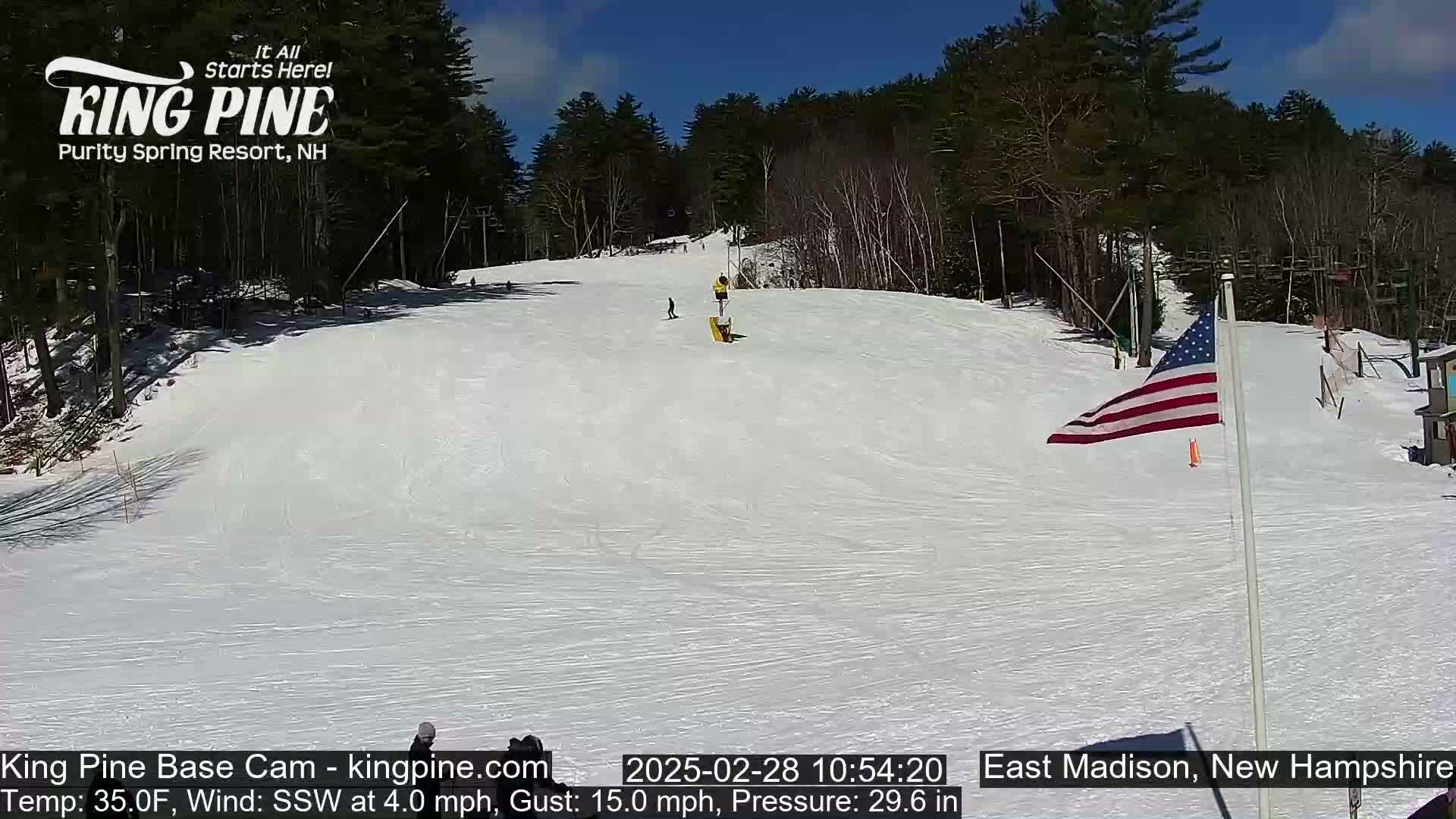 A snow-covered ski slope with several skiers and snowboarders, a US flag, and trees under a sunny sky.