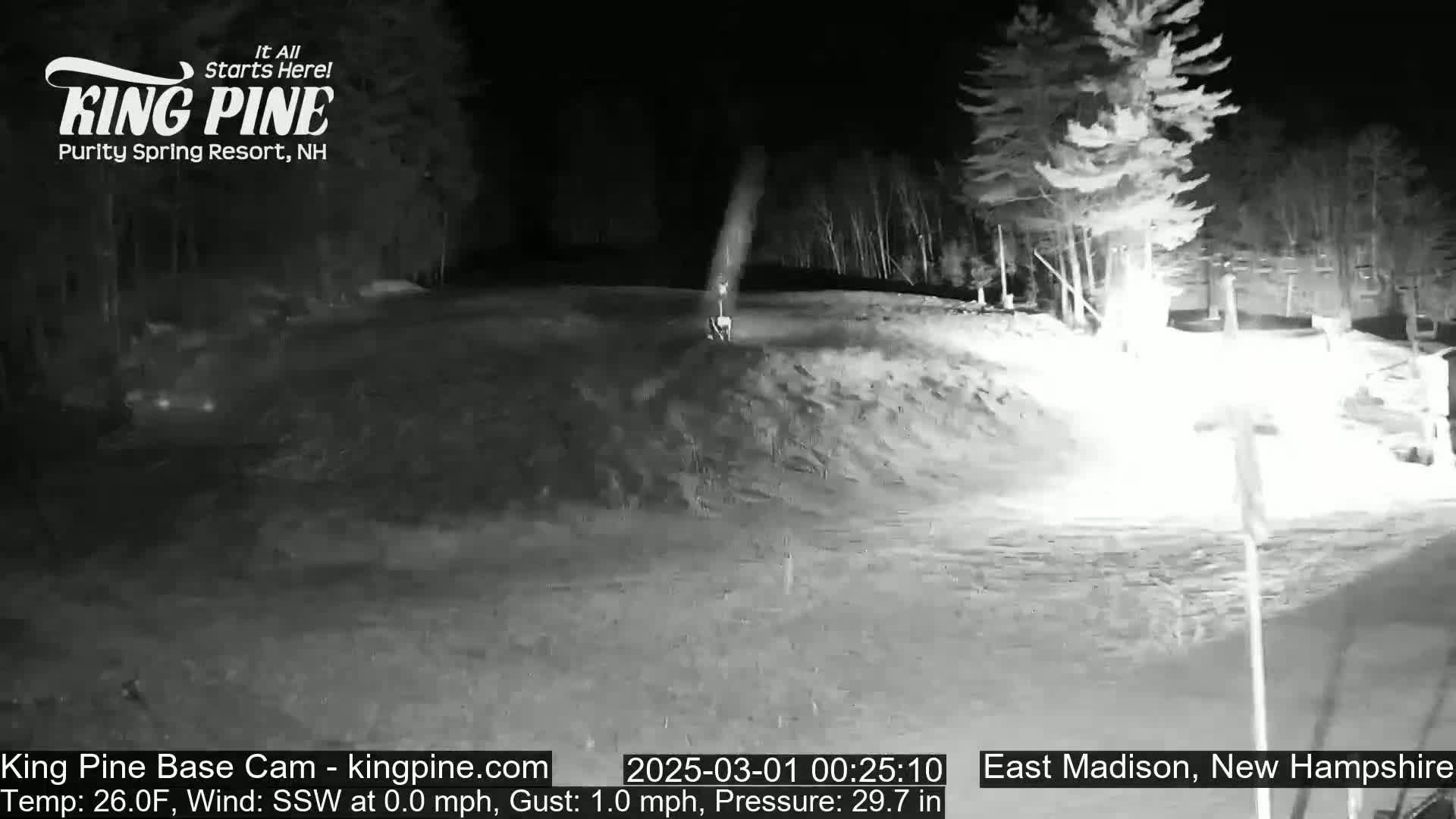 A nighttime, snow-covered scene shows a hill with trees in the background under a clear sky.