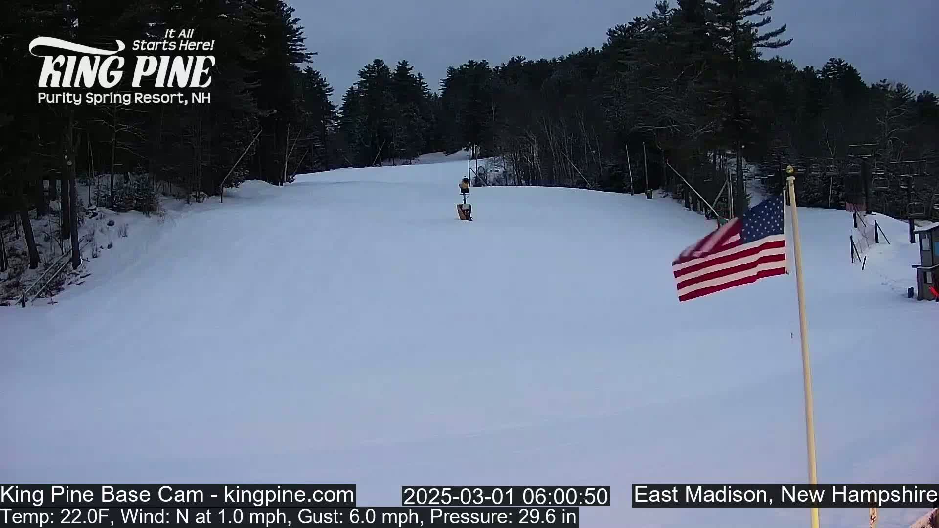 A snow-covered ski slope is seen under a cloudy, gray sky, with a small American flag waving gently in the breeze near a wooded area.