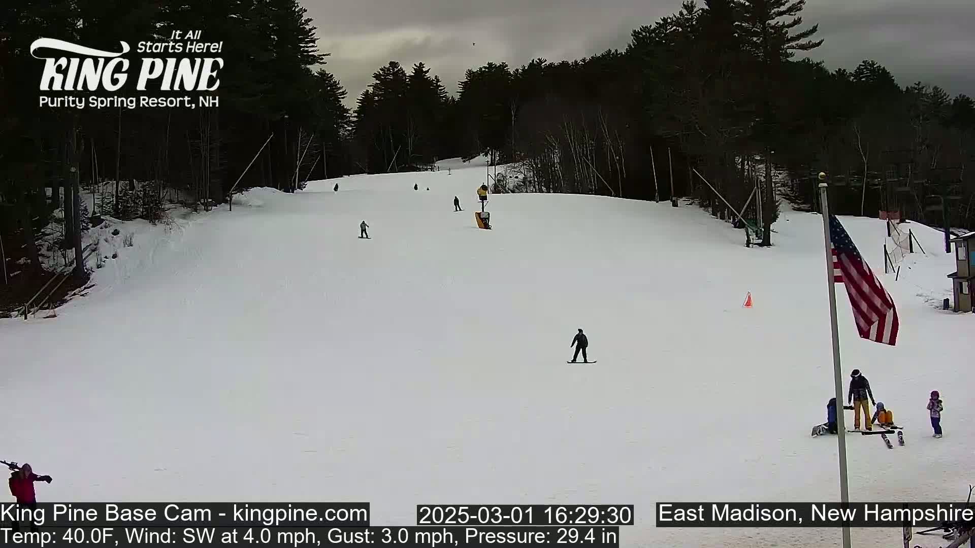 A snow-covered ski slope with several skiers and snowboarders, under an overcast sky, and a lightly blowing southwest wind.