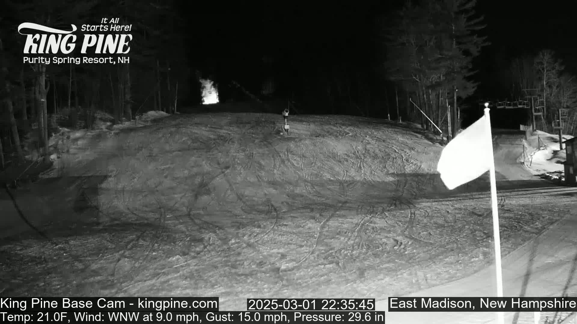 A nighttime, snow-covered ski slope with a lone figure and a white flag are visible under a WNW wind at 9.0 mph with gusts up to 15.0 mph and a temperature of 21°F.