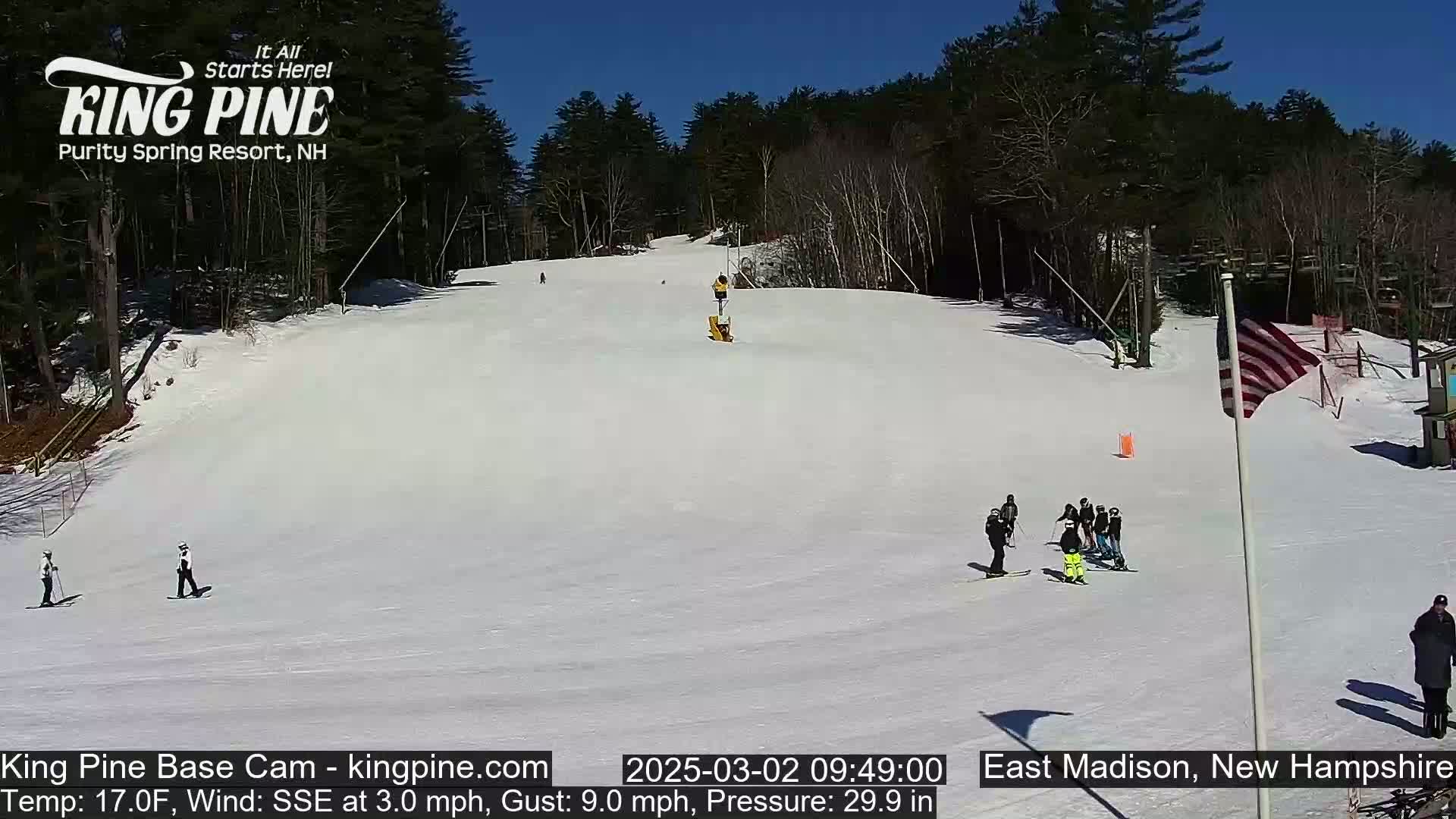 A snow-covered ski slope on a sunny day with several skiers and snowboarders visible, surrounded by trees.