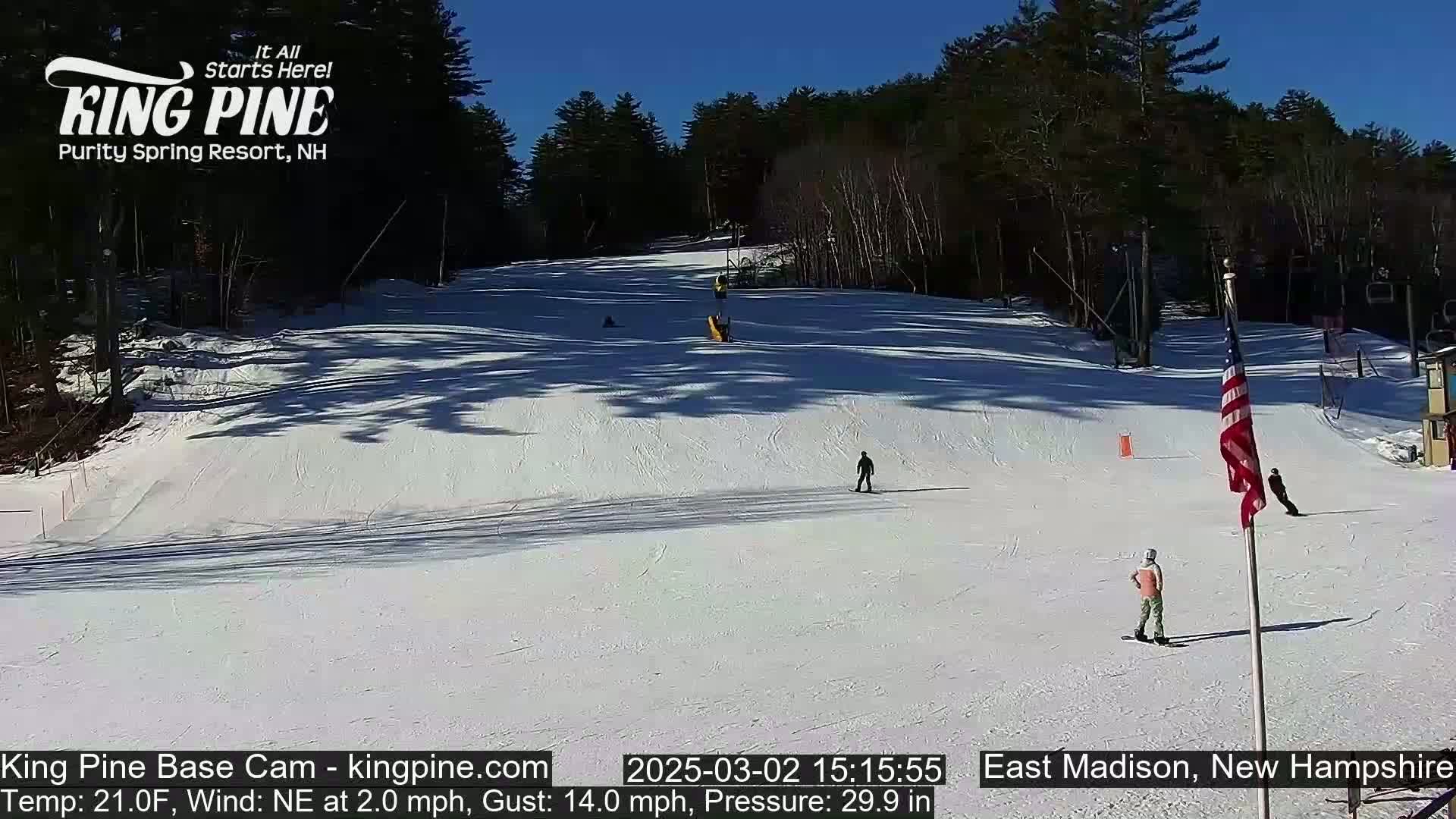 A snow-covered ski slope is visible on a sunny day with several skiers and snowboarders present.