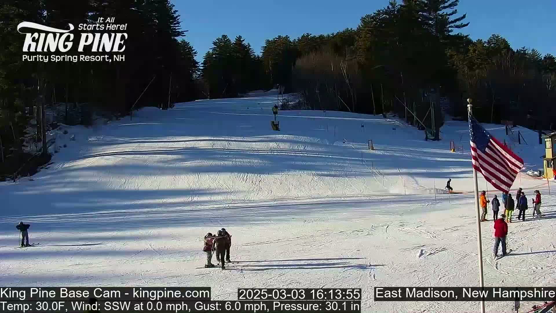 A snow-covered ski slope with several skiers and onlookers, under a clear blue sky.
