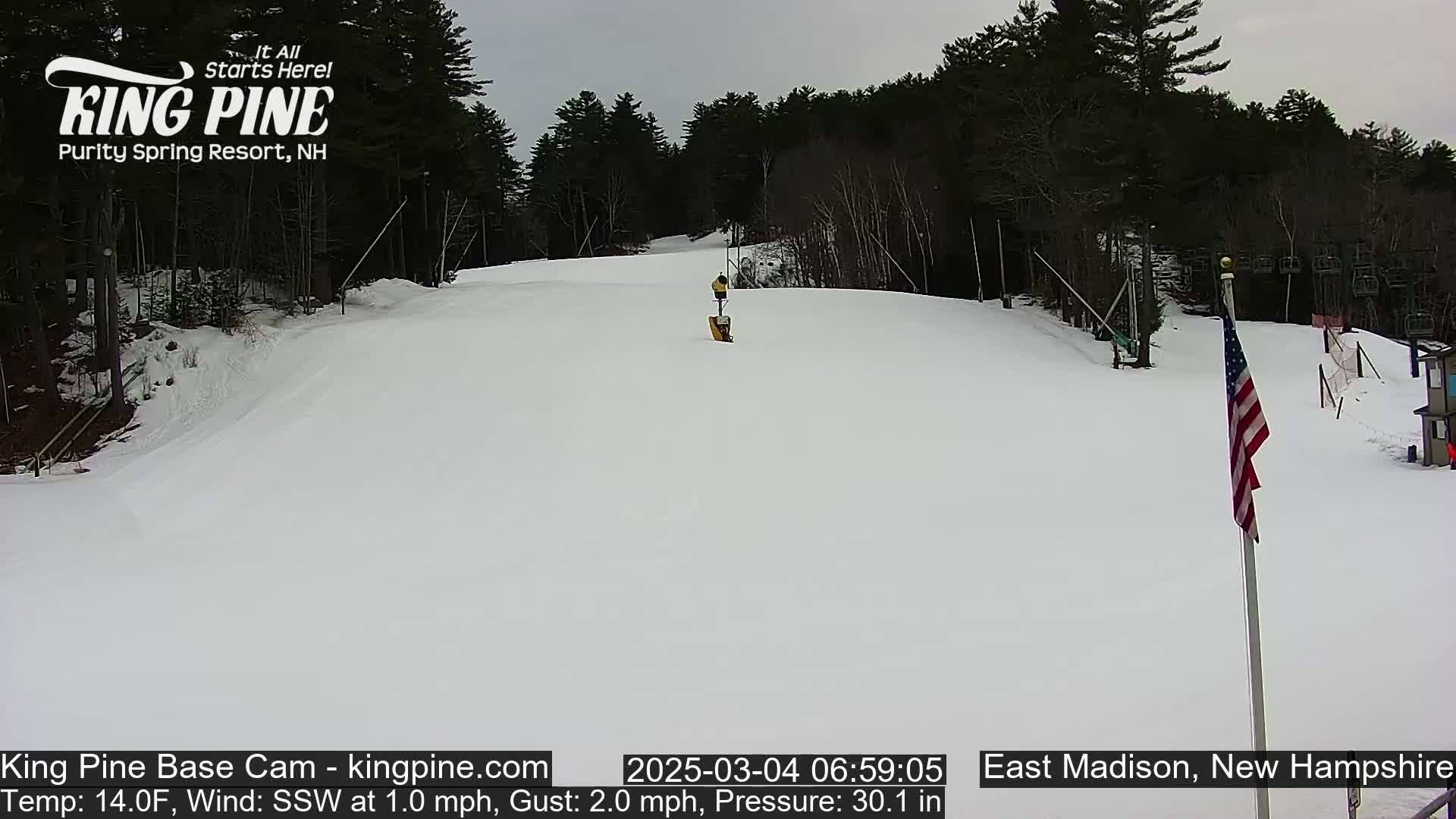 A snow-covered ski slope is visible, with a person on the slope and trees in the background under an overcast sky.