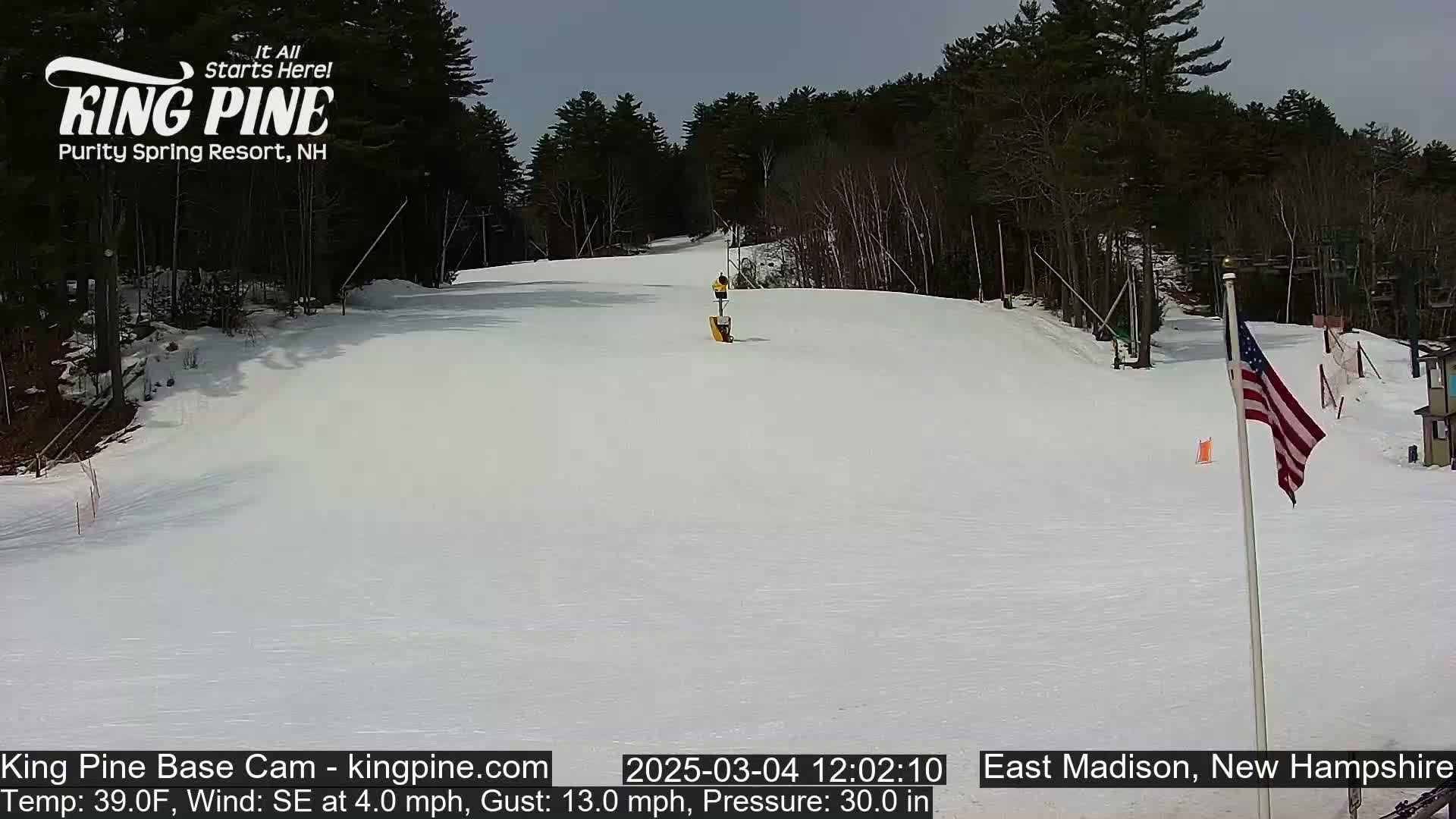 A snow-covered ski slope is visible under a mostly clear sky, with a few trees in the background and a small American flag in the foreground.
