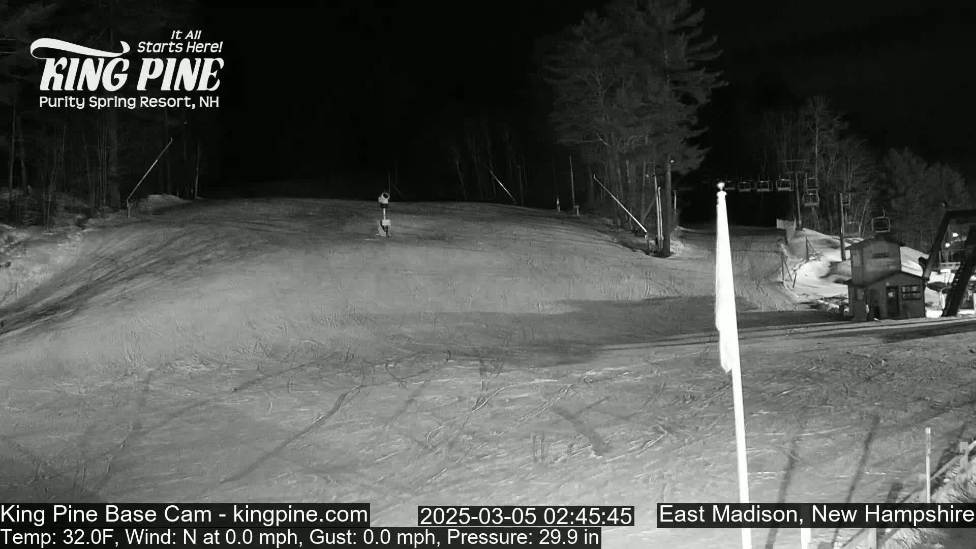 A snow-covered ski slope at night, illuminated by artificial lights, with a single person visible on the slope and a ski lift in the background.
