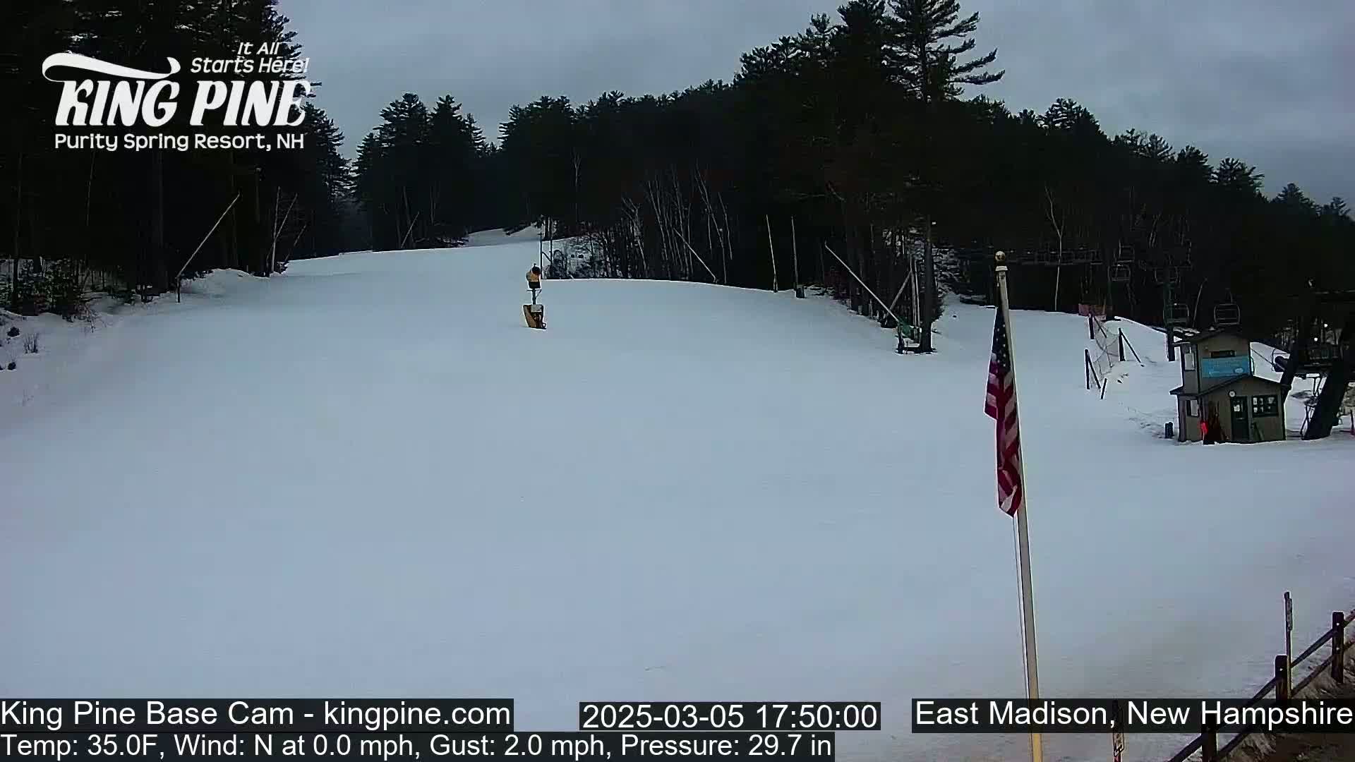 A snow-covered ski slope with a person skiing down it, under an overcast sky, is seen with a ski lift and a small building in the background.