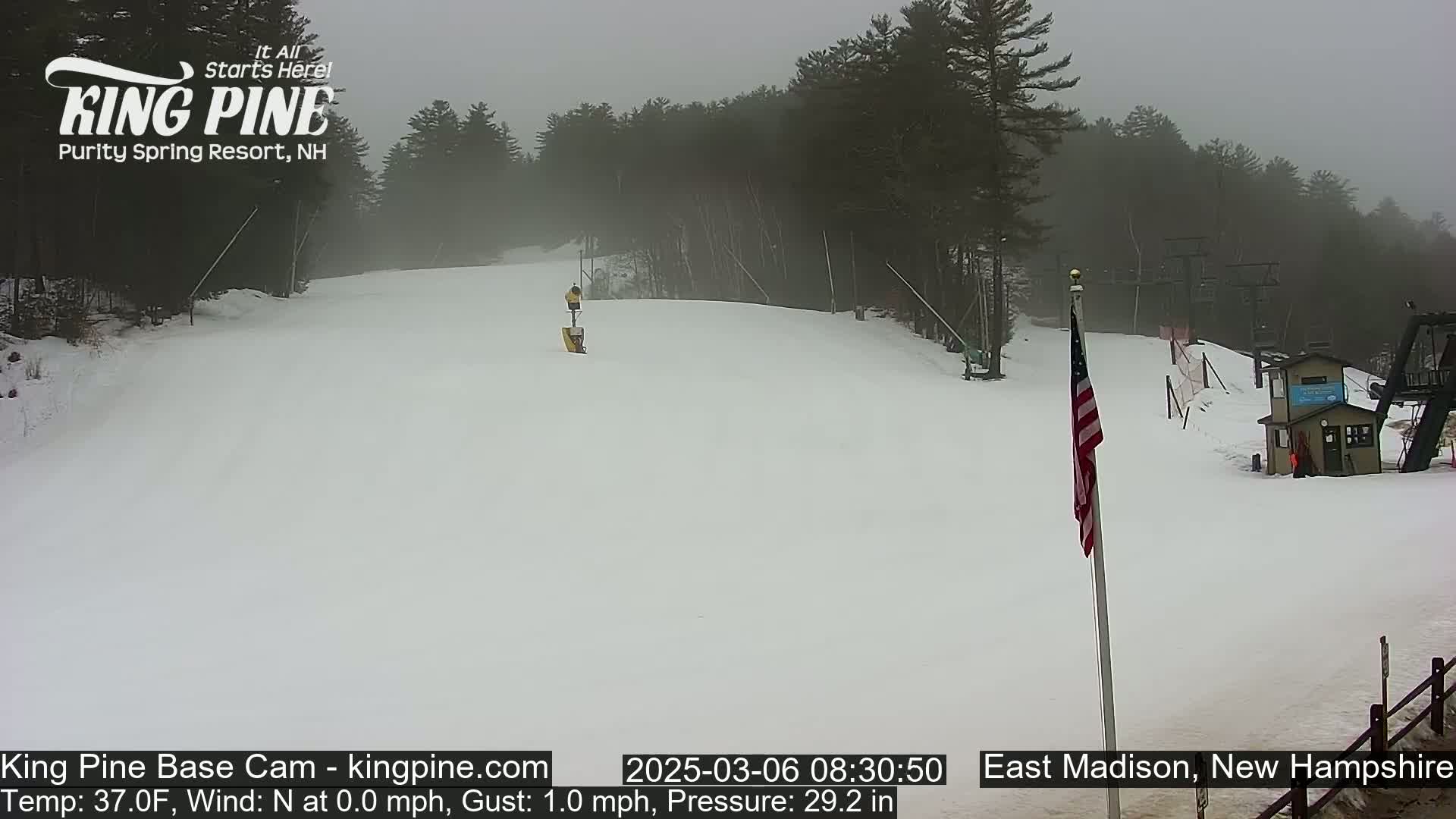 A snowy ski slope is visible under an overcast sky, with a person skiing down the hill and a small building in the background.