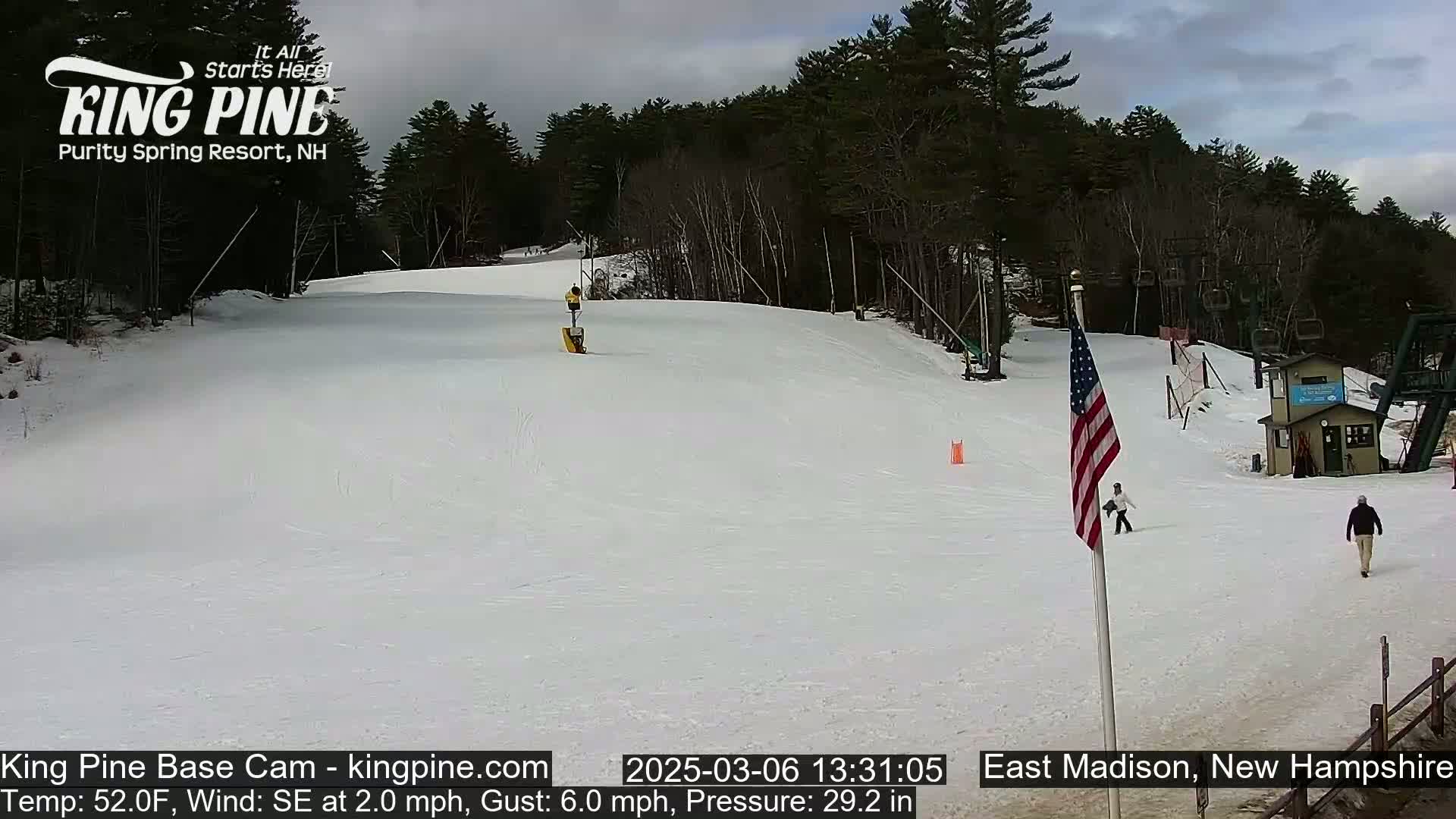 A snow-covered ski slope with a few skiers, a ski lift, a small building, and an American flag, under a cloudy sky.