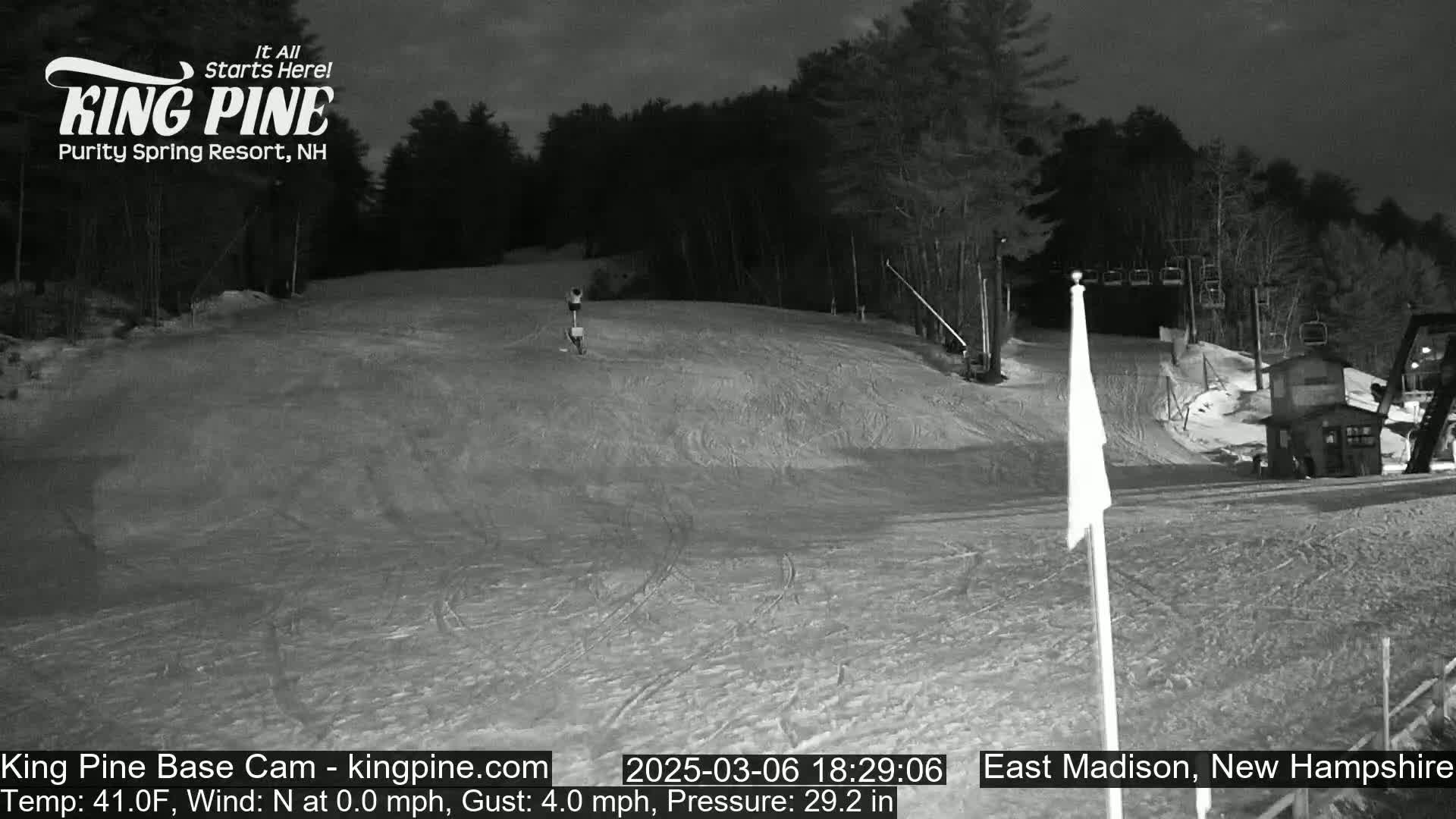 A nighttime, snow-covered ski slope with a ski lift and a small building in the background under a dark sky.