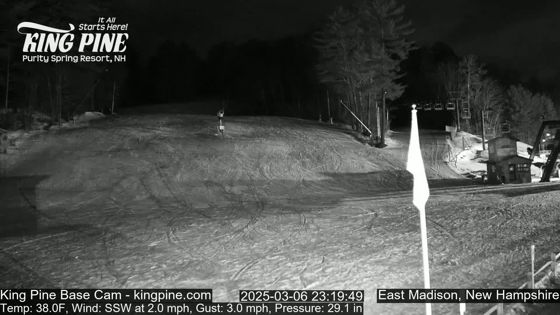 A nighttime, snow-covered ski slope is dimly lit, with a ski lift and trees in the background, under a clear sky.