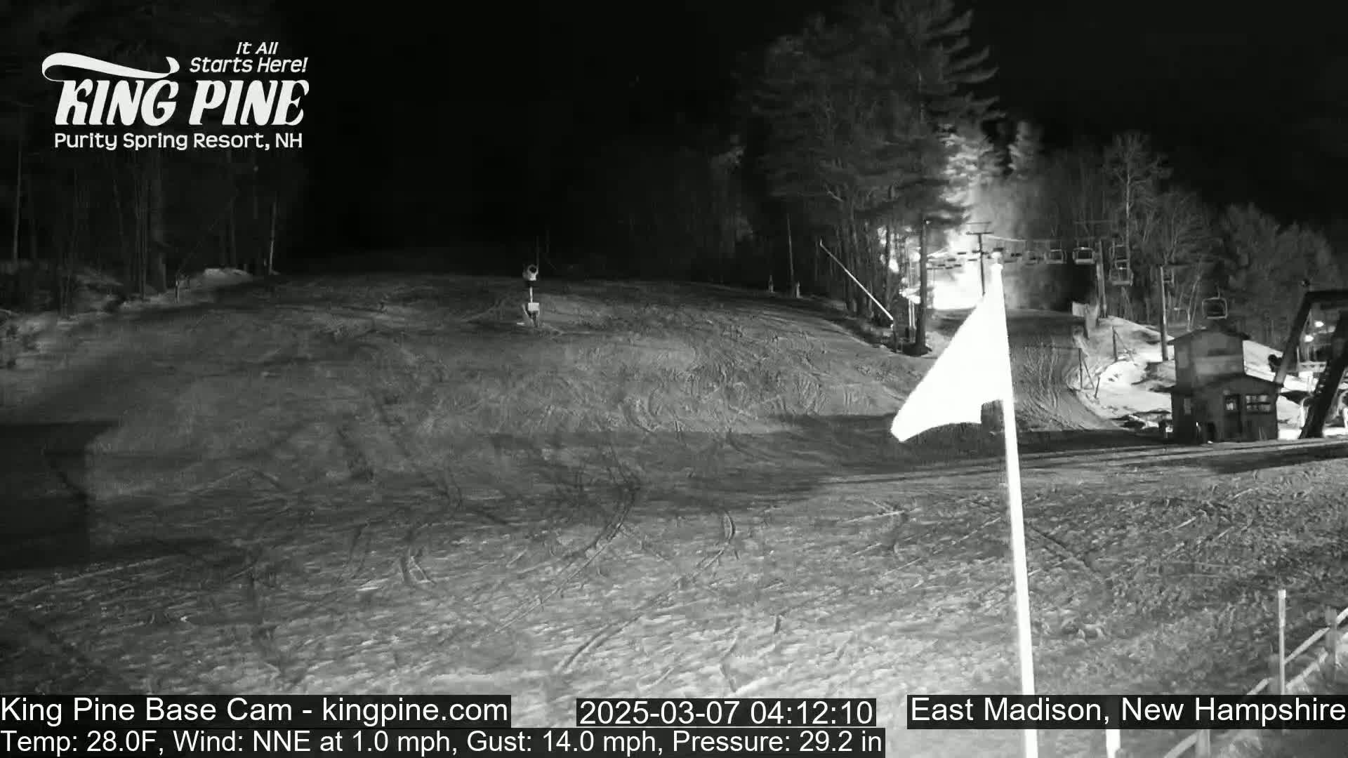 A nighttime view of a snow-covered ski slope with a ski lift and trees in the background under dark conditions.