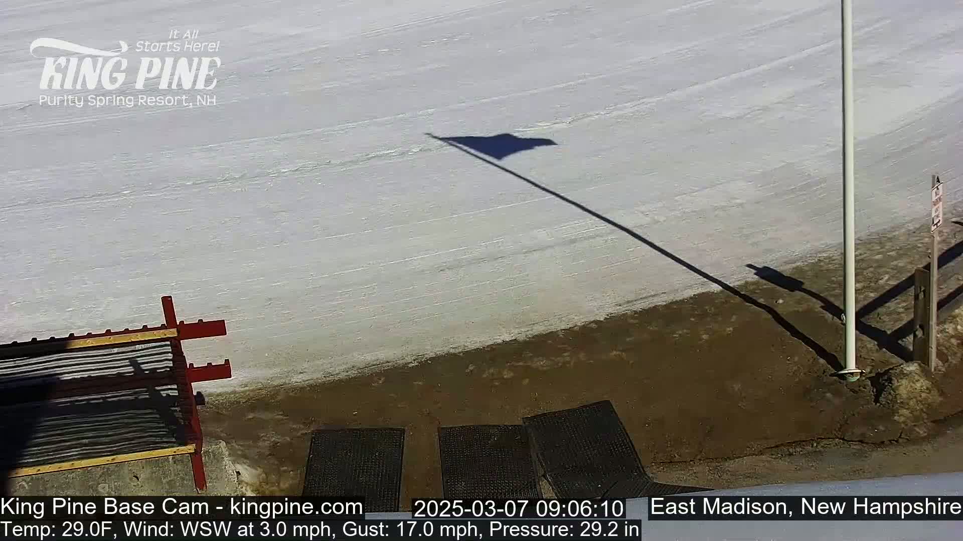 A snow-covered ski slope with a flag and its shadow cast on the ground, under a sunny sky with a slight wind.