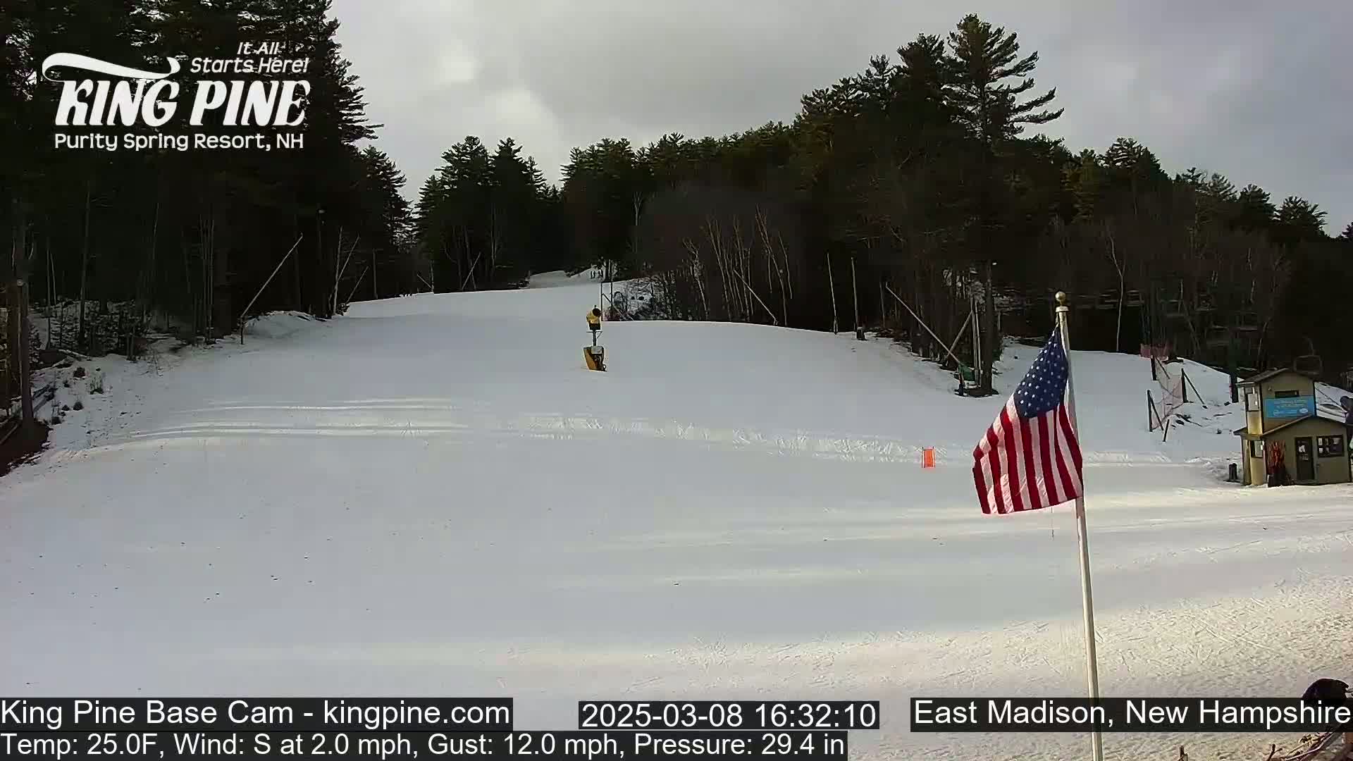 A snow-covered ski slope with a few skiers visible, under an overcast sky, is situated in a wooded area with an American flag in the foreground.