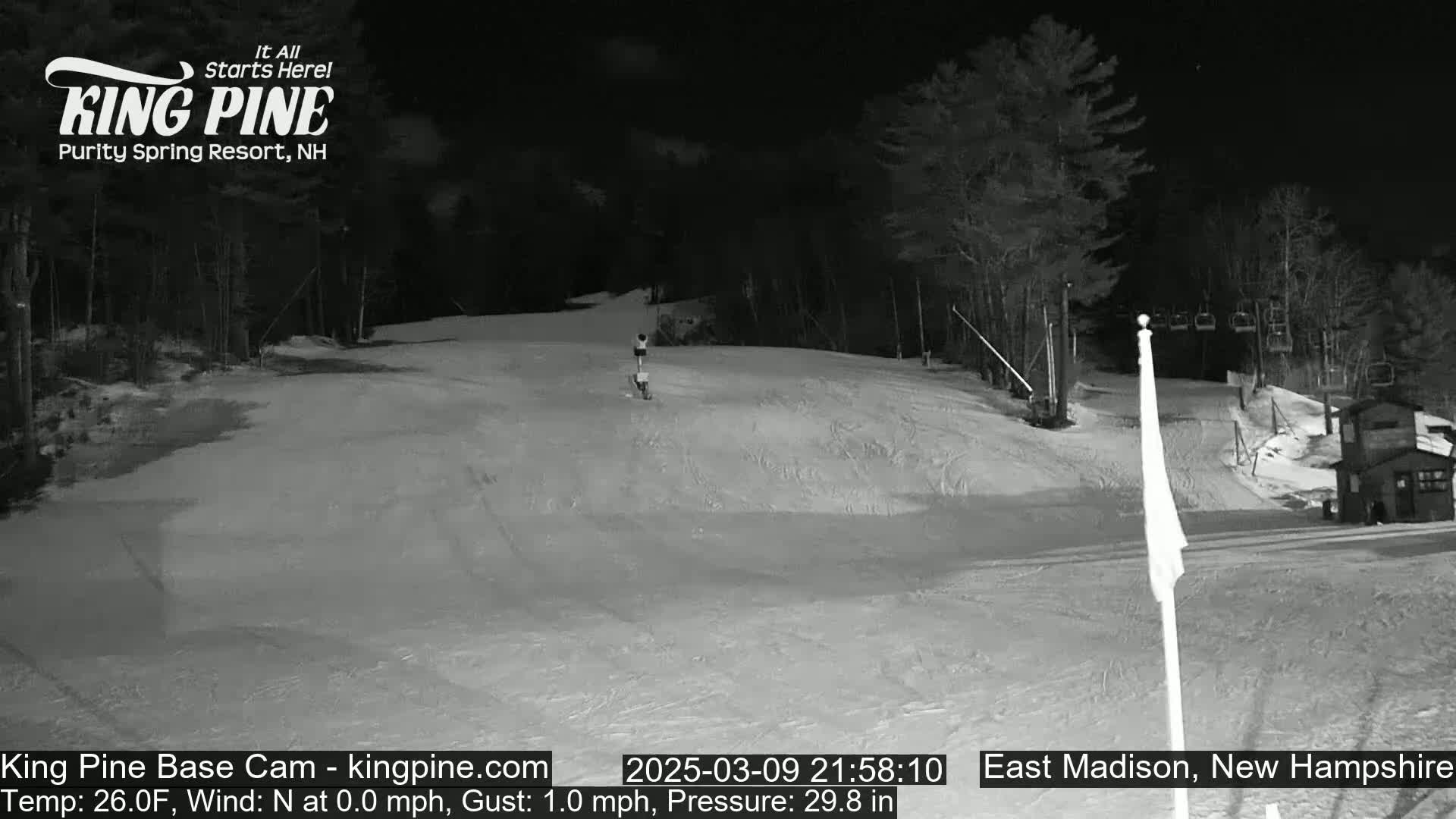 A nighttime view of a snow-covered ski slope with trees in the background under a clear, dark sky.